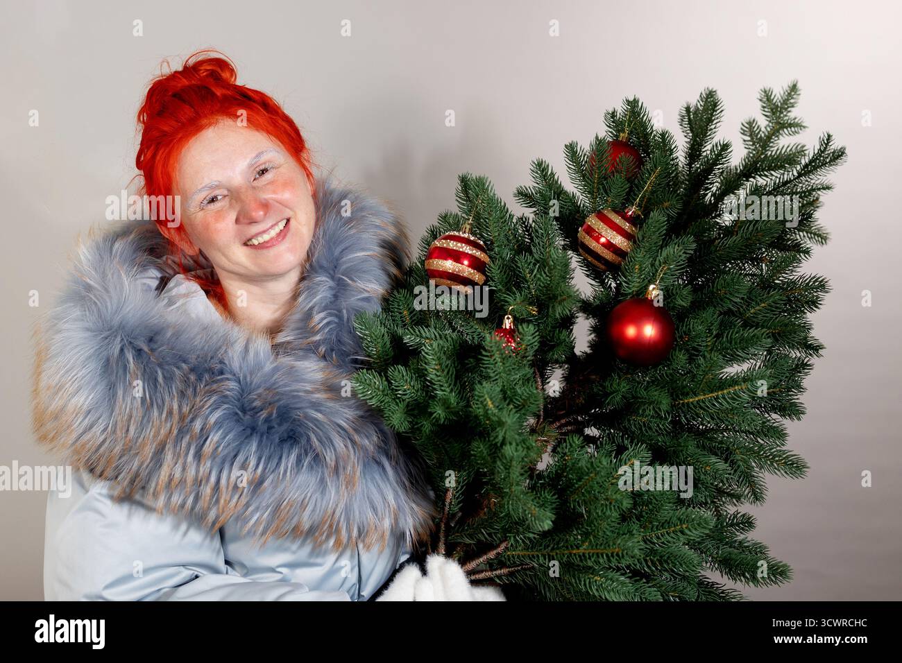 Frau im Wintermantel lächelt, nachdem sie draußen in der Kälte war. Sie hält einen kleinen grünen Weihnachtsbaum, der mit roten und goldenen Ornamenten verziert ist. Stockfoto