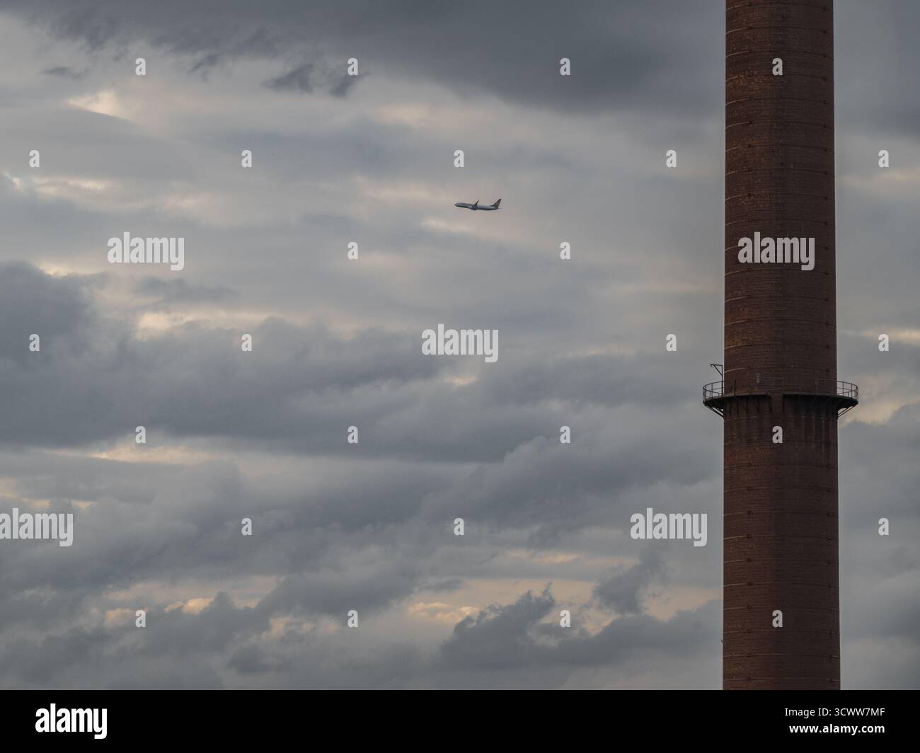 Das Flugzeug fliegt an industriellem Kamin unter dramatischem bewölktem Himmel vorbei und symbolisiert Reisen und Industrie Stockfoto