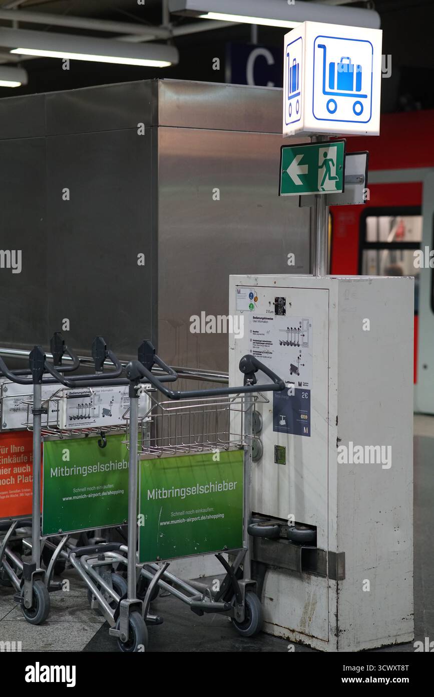 Gepäckwagen und Rückkehrkasse im Münchner Flughafenterminal mit beleuchteten Schildern und Reisesymbolen. München, Deutschland Stockfoto