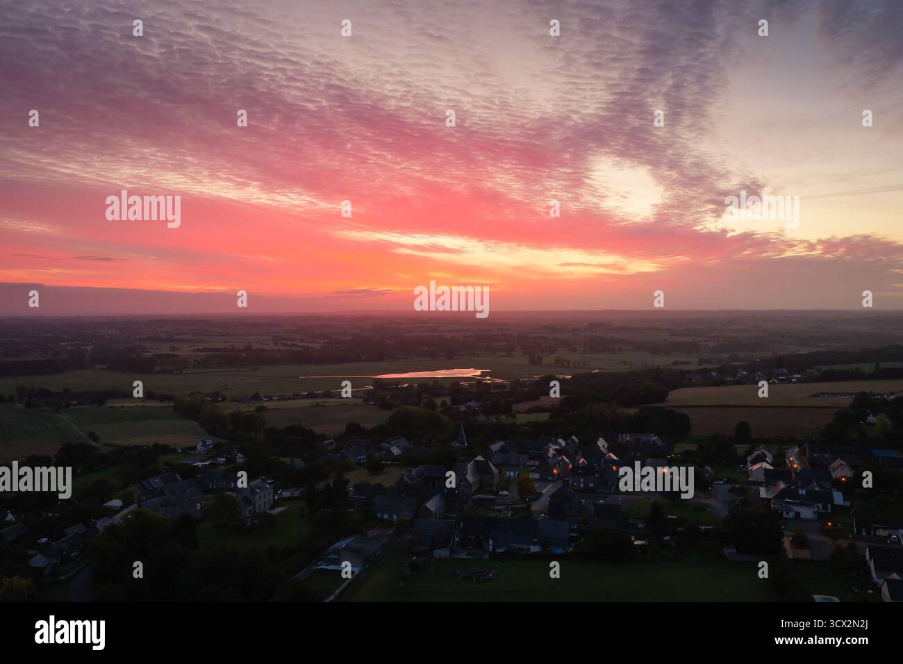 Blick aus der Vogelperspektive auf ein kleines ländliches Dorf bei Sonnenuntergang. Der Himmel leuchtet rosa und orange und reflektiert auf Feldern und einem fernen Wasser. Ruhig, landschaftlich und ideal Stockfoto