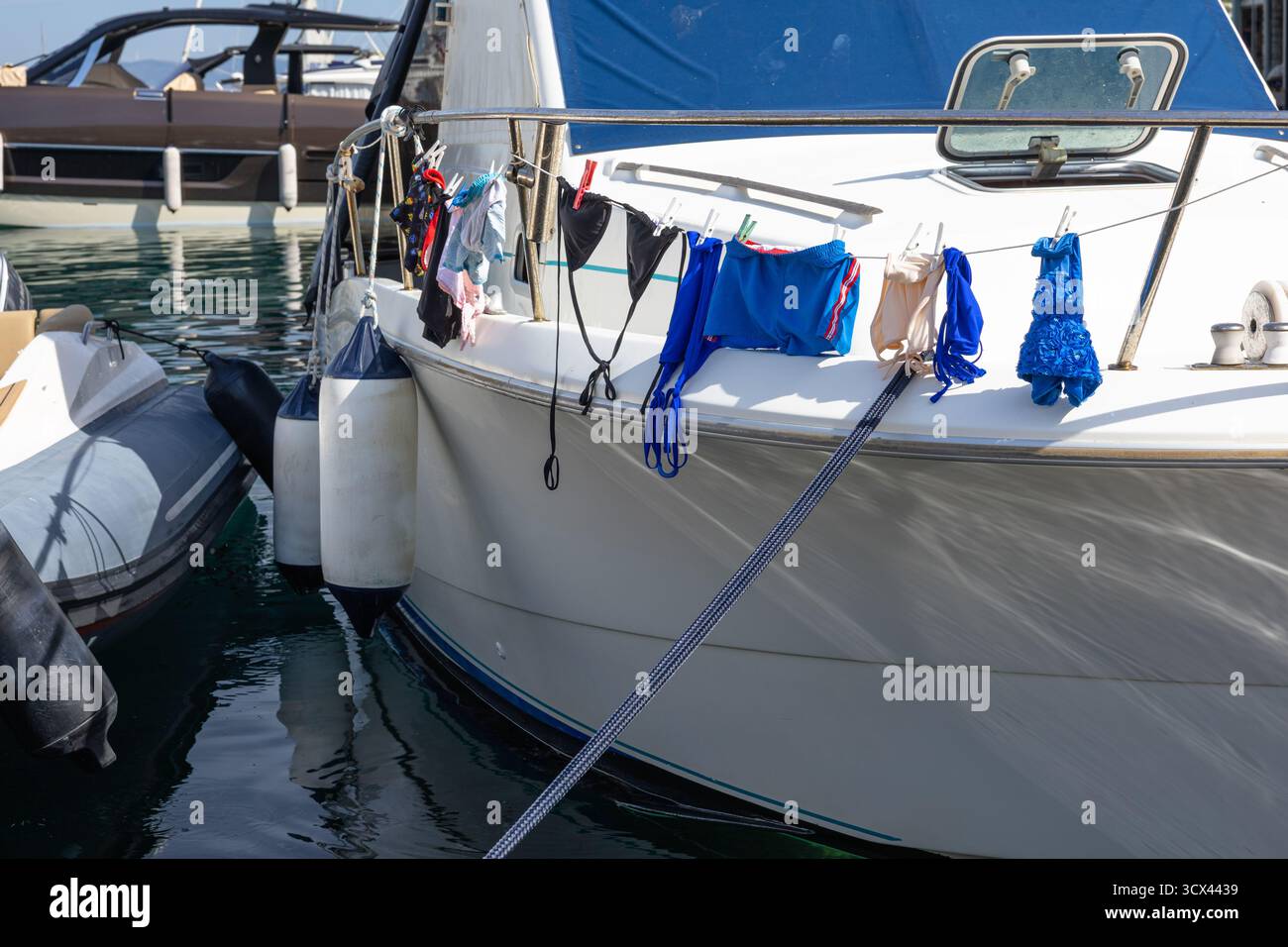 Ein Segelboot, das an einem sonnigen Yachthafen anlegt, zeigt bunte Kleidung, die entlang der Schiene trocknet, mit Fdern und Festmacherleinen, die das Schiff sichern. Ruhiges Wasser und n Stockfoto