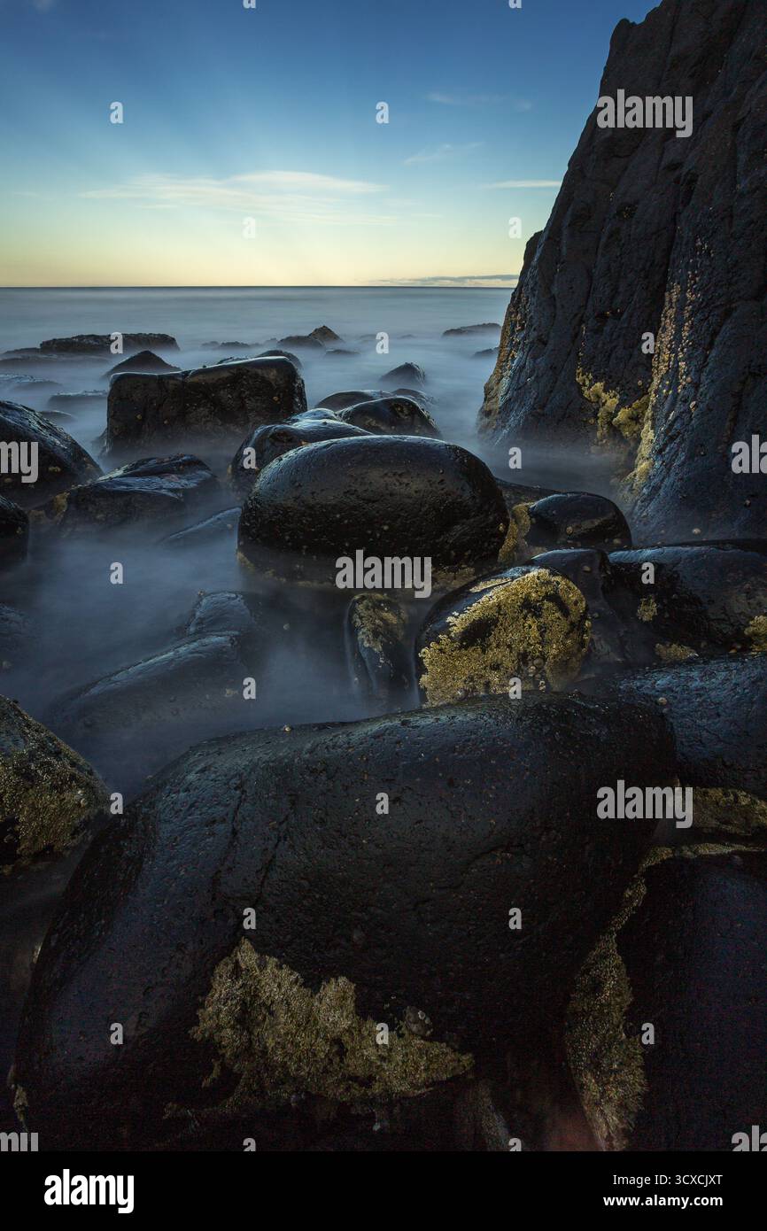 Felsige Küste in Burleigh Heads, Australien. Große, glatte, dunkle Felsbrocken werden teilweise in nebeliges Wasser getaucht, das während des Sonnenaufgangs gefangen wird. Stockfoto