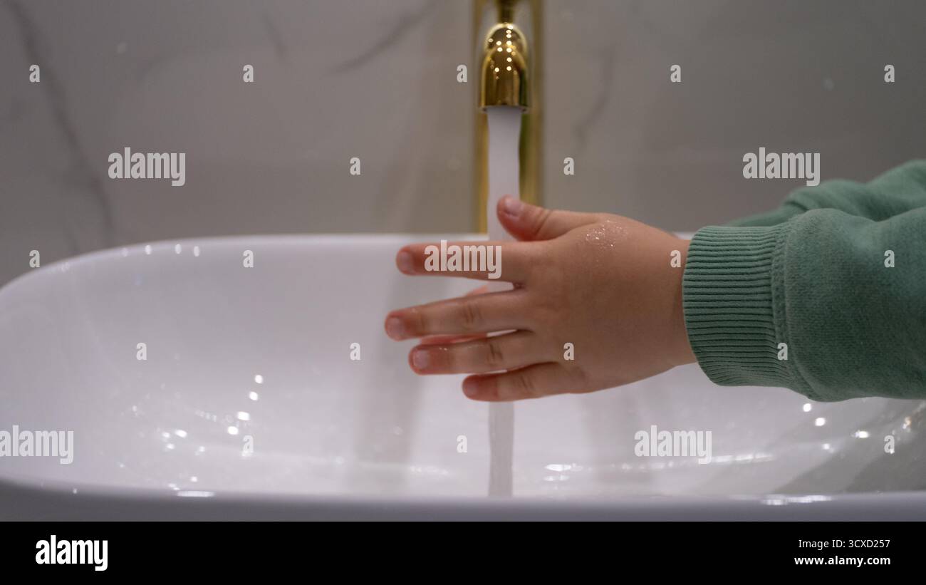Minimalistisches Bild einer Kinderhand unter fließendem Wasser eines goldenen Wasserhahns. Symbolische Darstellung der täglichen Hygiene, Sauberkeit und Gesundheitserziehung Stockfoto