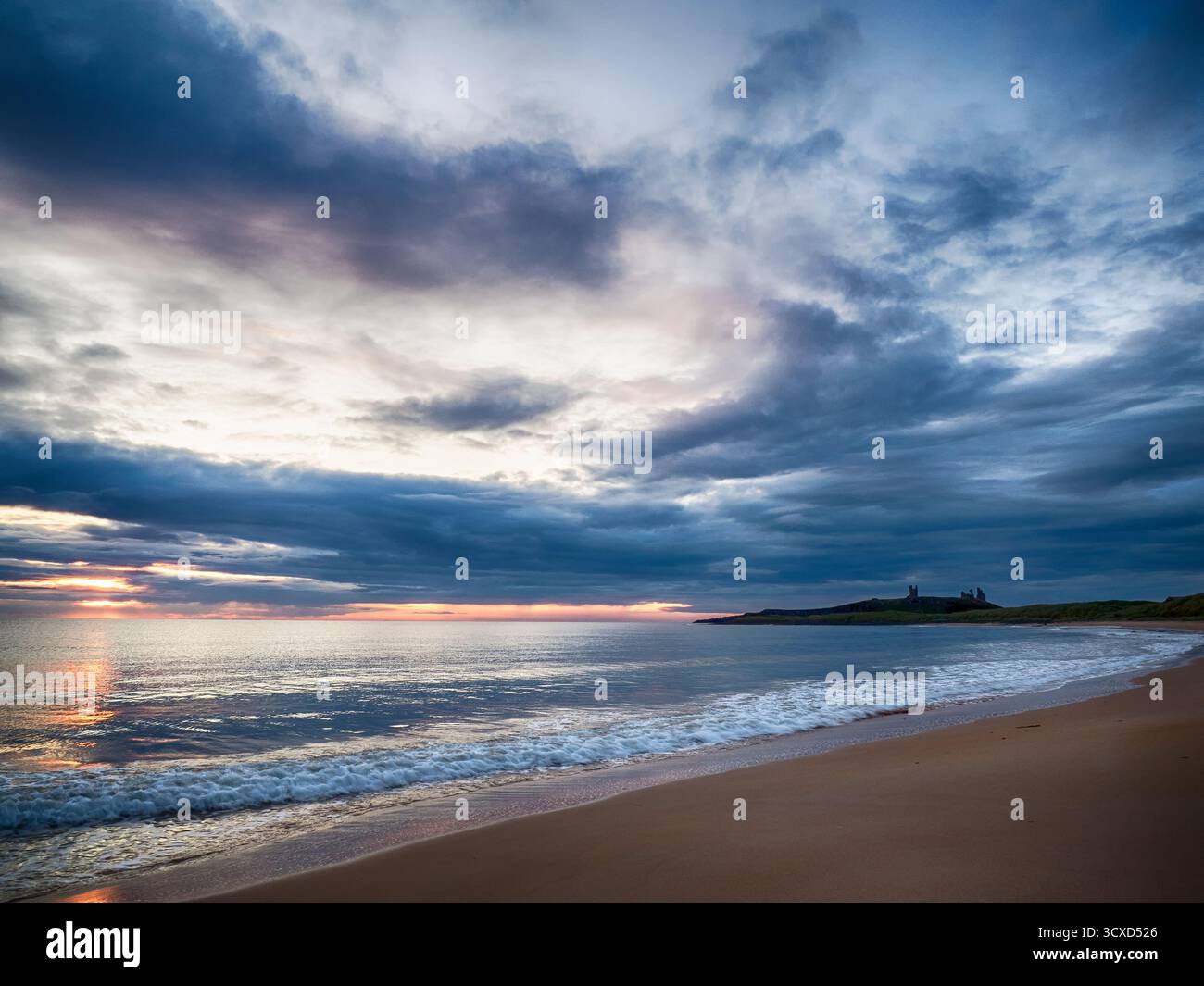 Dunstanburgh Castle bei Sonnenaufgang vom Strand aus mit sanften Wellen unter einem stimmungsvollen Himmel Stockfoto