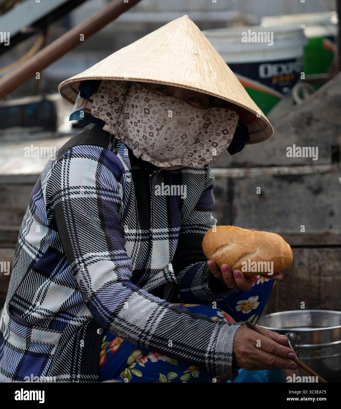 Frau mit konischem Hut und Gesichtsbedeckung, die einen Brotlaib auf einem schwimmenden Markt in Vietnam hält Stockfoto