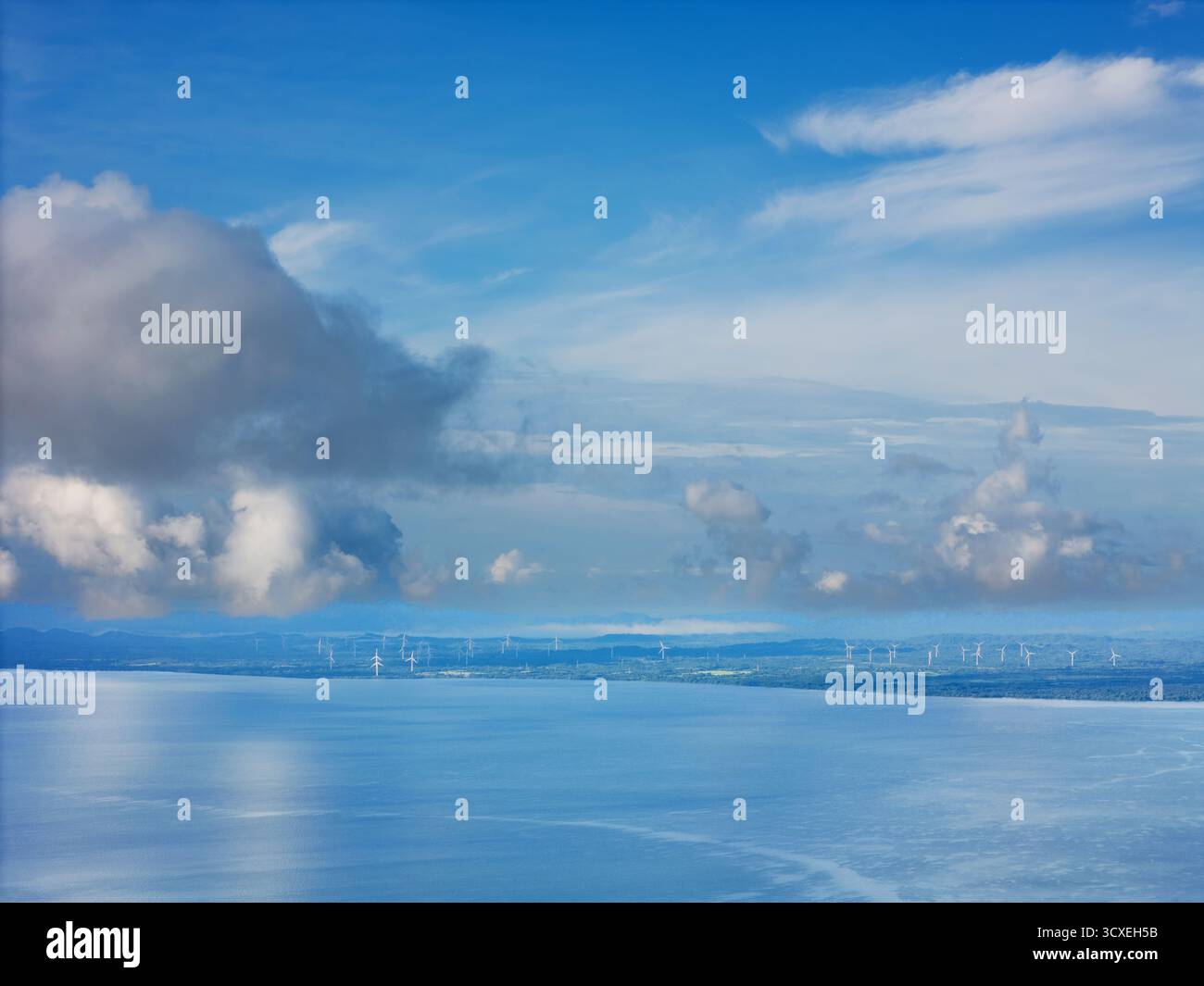 Windturbinen säumen die ferne Küste, während die Wellen sanft am Ufer unter dem ruhigen blauen Himmel liegen. Stockfoto