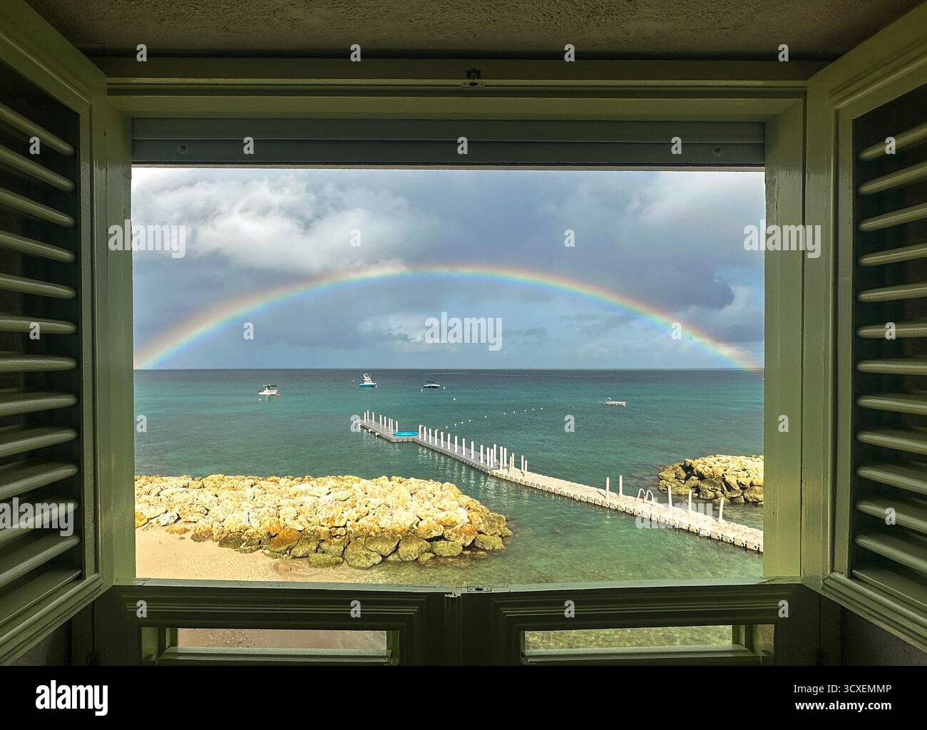 Blick eingerahmt durch ein offenes Hotelfenster mit Rollläden zum Barbados Beach, zum türkisfarbenen Meer, zu Booten und zum Bootsanleger am Sommertag mit Rainbow Stockfoto