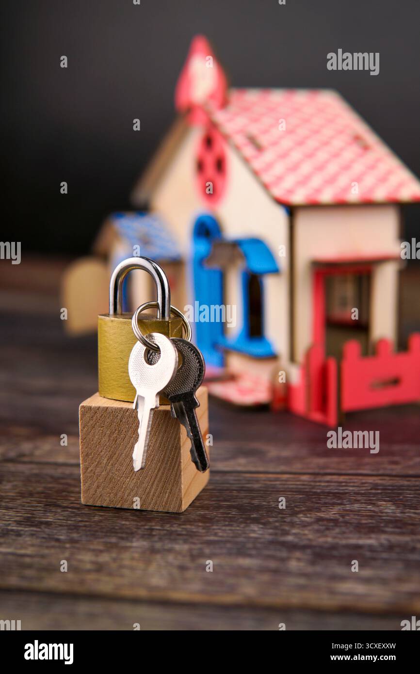 Vorhängeschloss und Schlüssel auf einem Holzblock vor einem verschwommenen Miniaturhaus, das Wohneigentum, finanzielle Sicherheit, Immobilieninvestition repräsentiert. Stockfoto