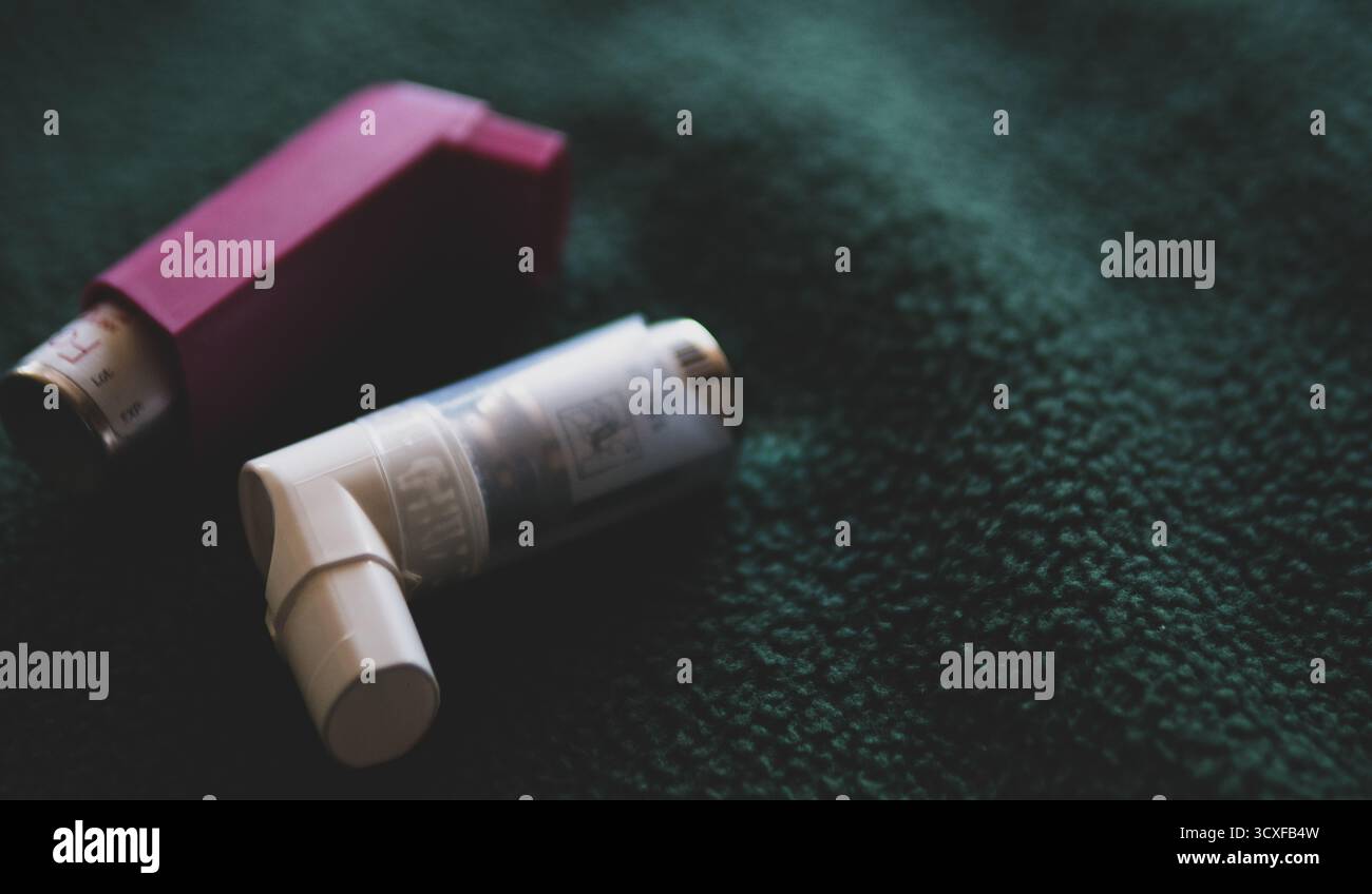 Nahaufnahme von zwei Asthma-Inhalatoren, einer rosa und einer weiß, auf einer dunkelgrünen texturierten Stoffoberfläche platziert.symbolisiert Gesundheit, Medizin. Stockfoto