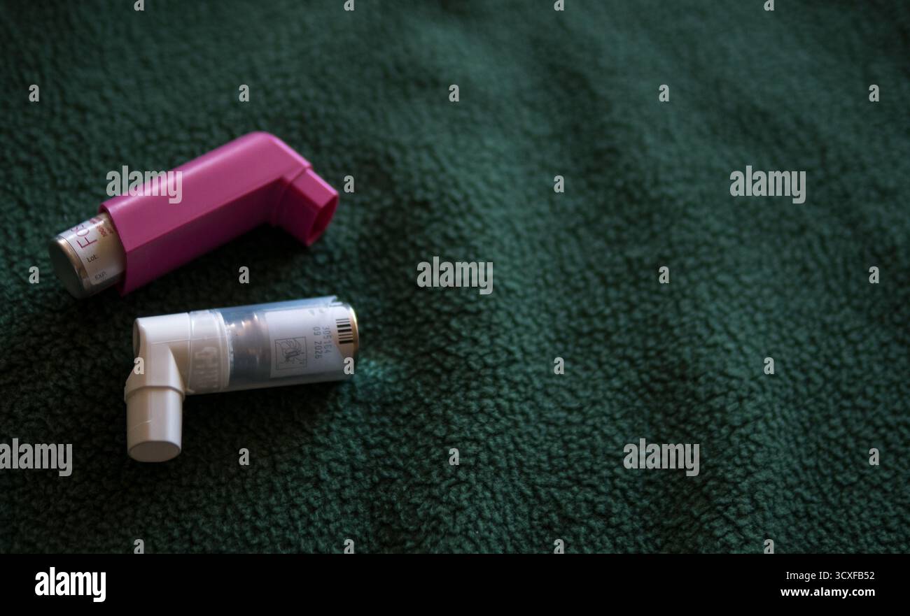 Nahaufnahme von zwei Asthma-Inhalatoren, einer rosa und einer weiß, auf einer dunkelgrünen texturierten Stoffoberfläche platziert.symbolisiert Gesundheit, Medizin. Stockfoto