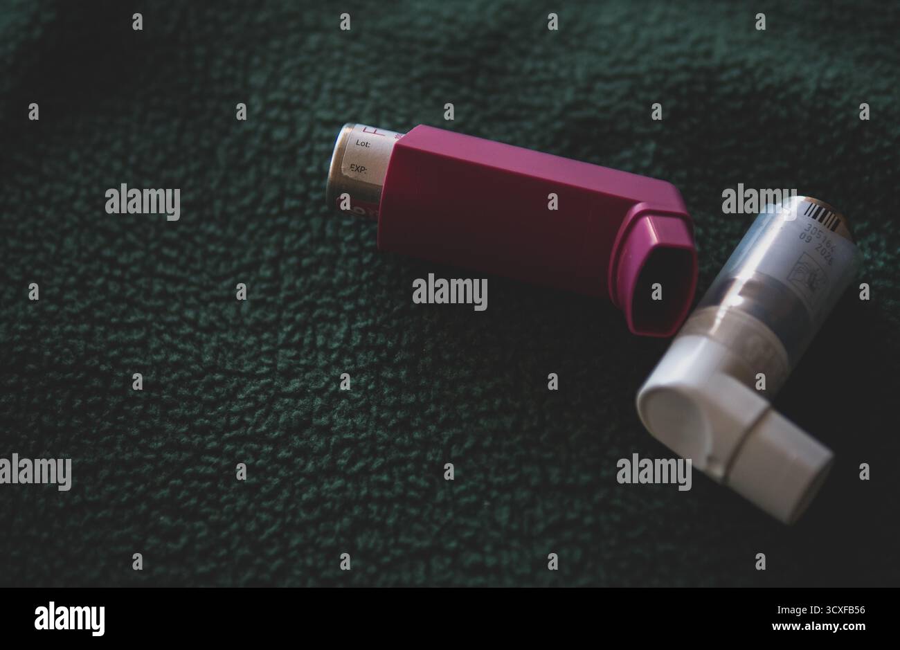 Nahaufnahme von zwei Asthma-Inhalatoren, einer rosa und einer weiß, auf einer dunkelgrünen texturierten Stoffoberfläche platziert.symbolisiert Gesundheit, Medizin. Stockfoto