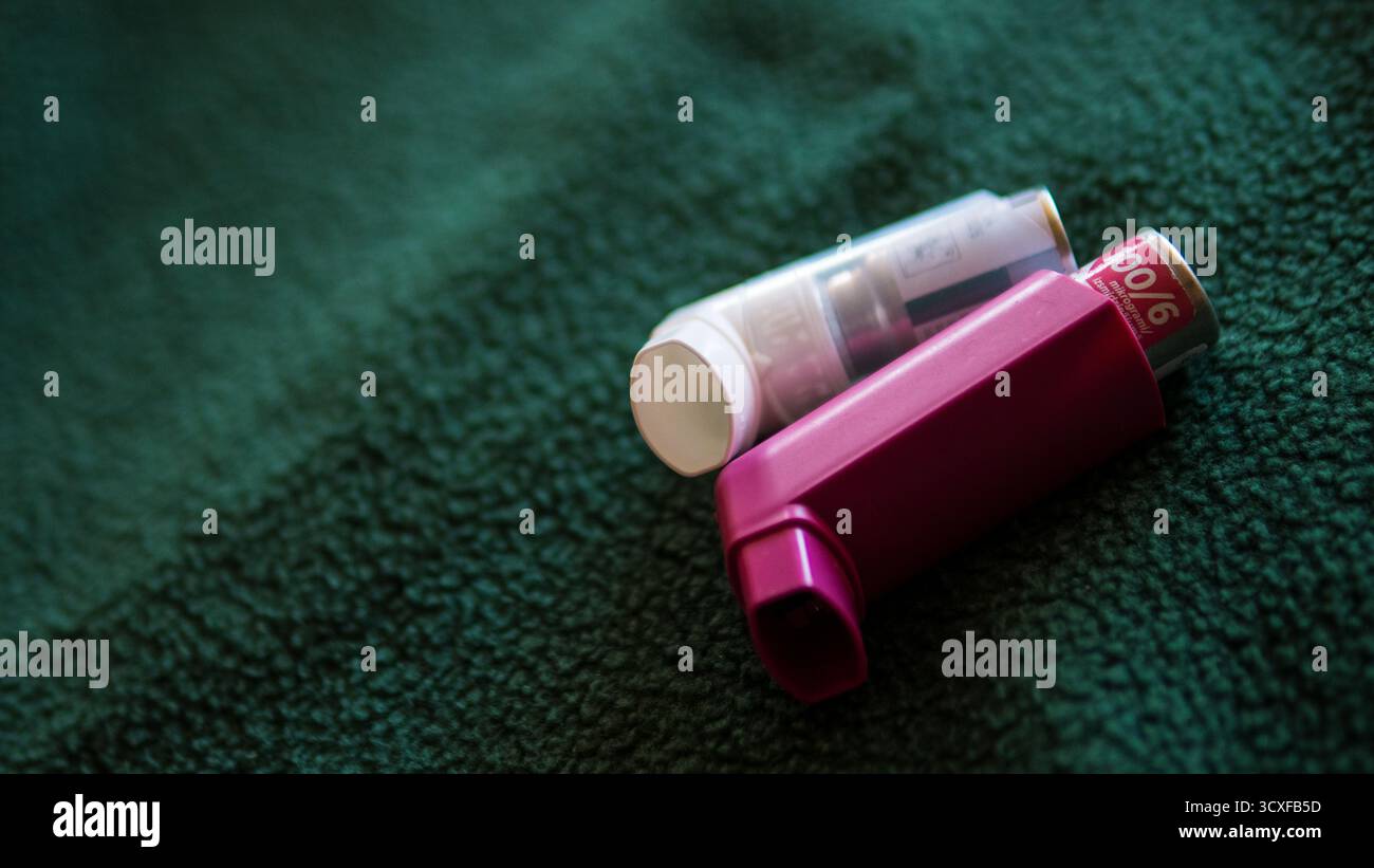 Nahaufnahme von zwei Asthma-Inhalatoren, einer rosa und einer weiß, auf einer dunkelgrünen texturierten Stoffoberfläche platziert.symbolisiert Gesundheit, Medizin. Stockfoto