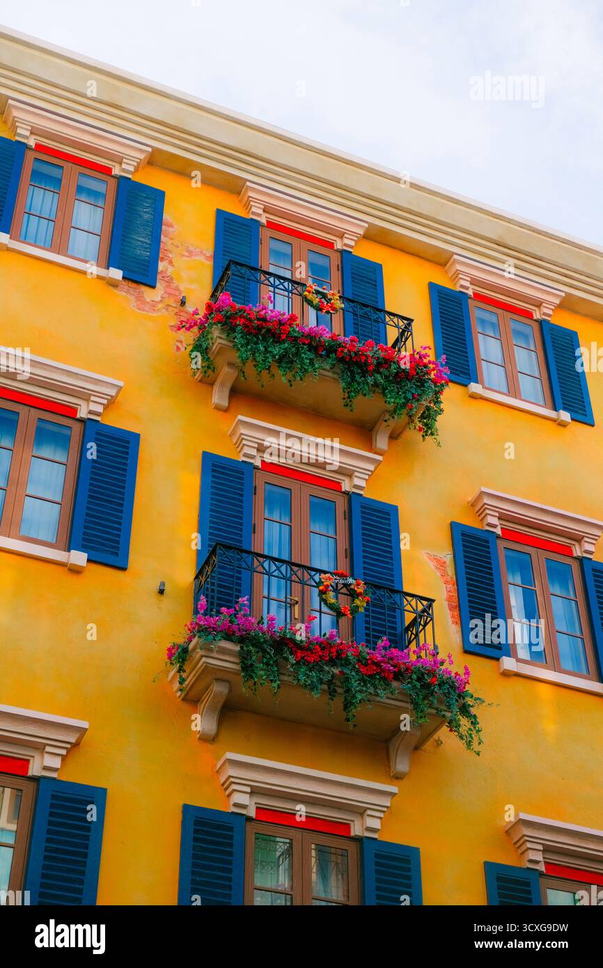 Farbenfrohe Fassade im mediterranen Stil mit blauen Fensterläden, roten Fensterrahmen und lebendigen Balkonen mit Blumendecken vor sonnigem Himmel Stockfoto