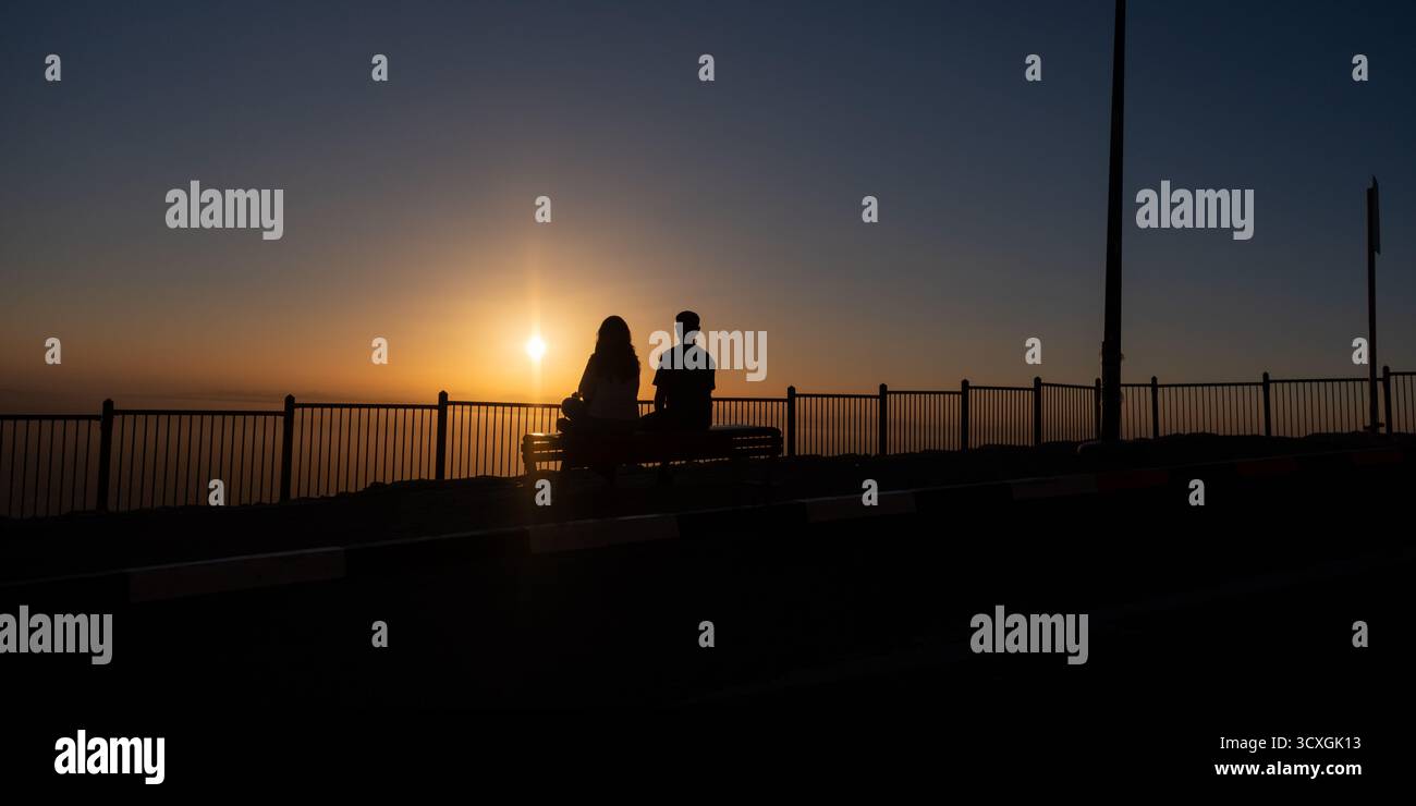 Silhouette eines Paares, das am Ufer sitzt und den Sonnenuntergang über dem Meer beobachtet Stockfoto