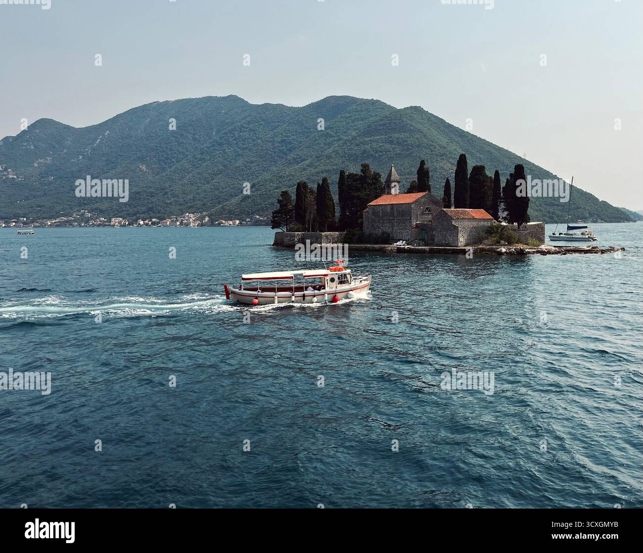 Ein kleines Passagierboot fährt an der Insel unserer Lieben Frau von den Felsen in der Bucht von Kotor vorbei, umgeben von grünen Bergen und ruhigen Adria-Gewässern. - Smartphone-aufgenommenes Stockfoto