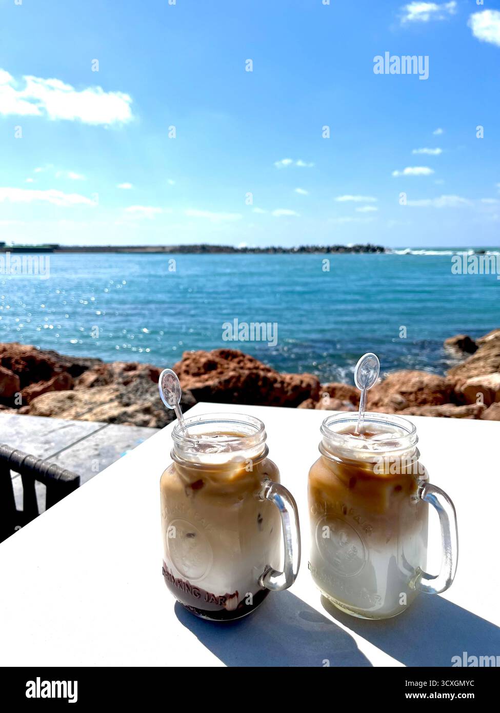 Erfrischende zwei Eiskaffeetassen am Meer an einem hellen Sommertag mit Blick auf das ruhige blaue Wasser. - Smartphone-aufgenommenes Stockfoto
