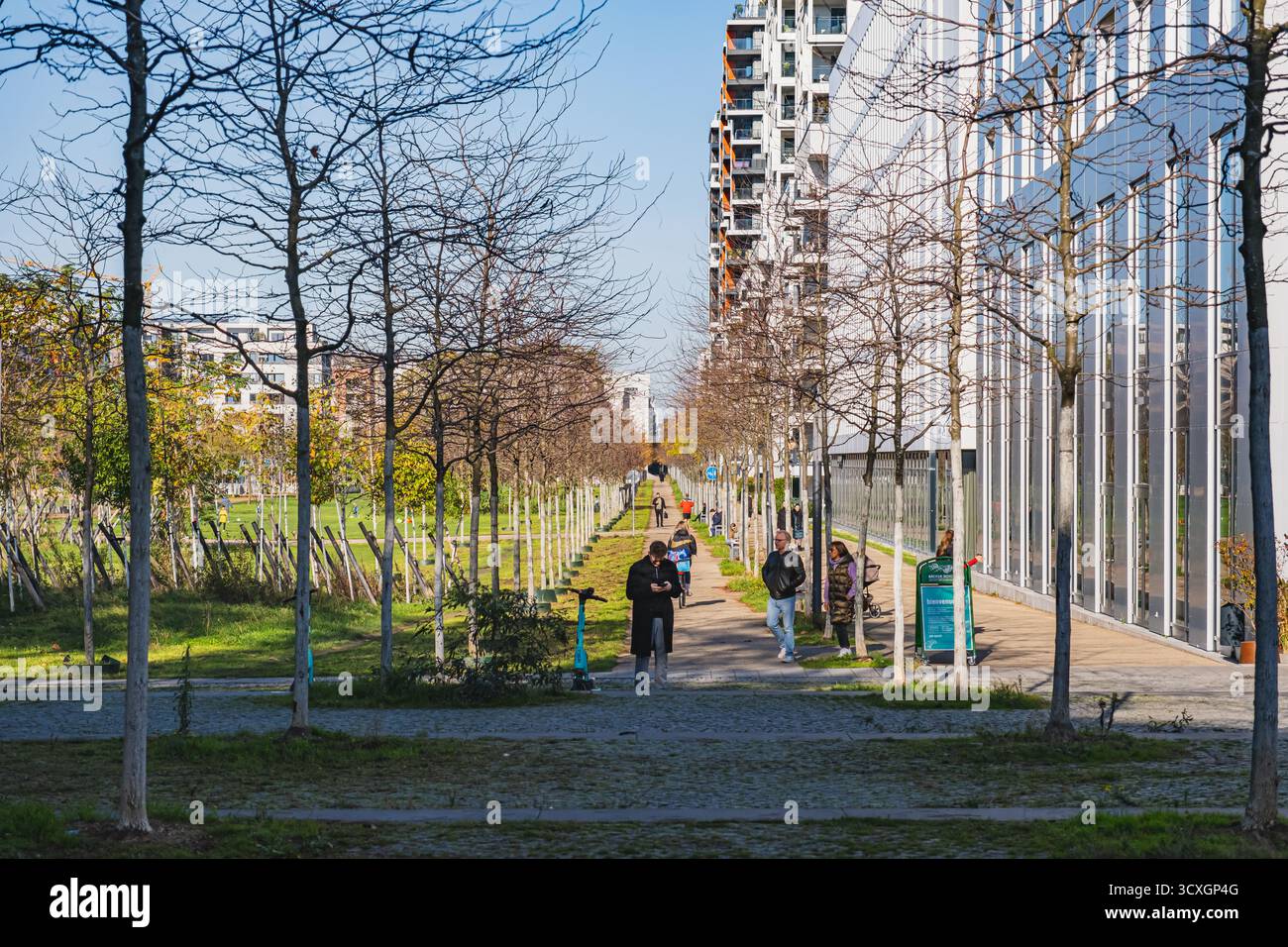 Fußgänger- und Radweg führt entlang moderner Wohngebäude, mit klarem Himmel und einem Park auf der linken Seite Stockfoto