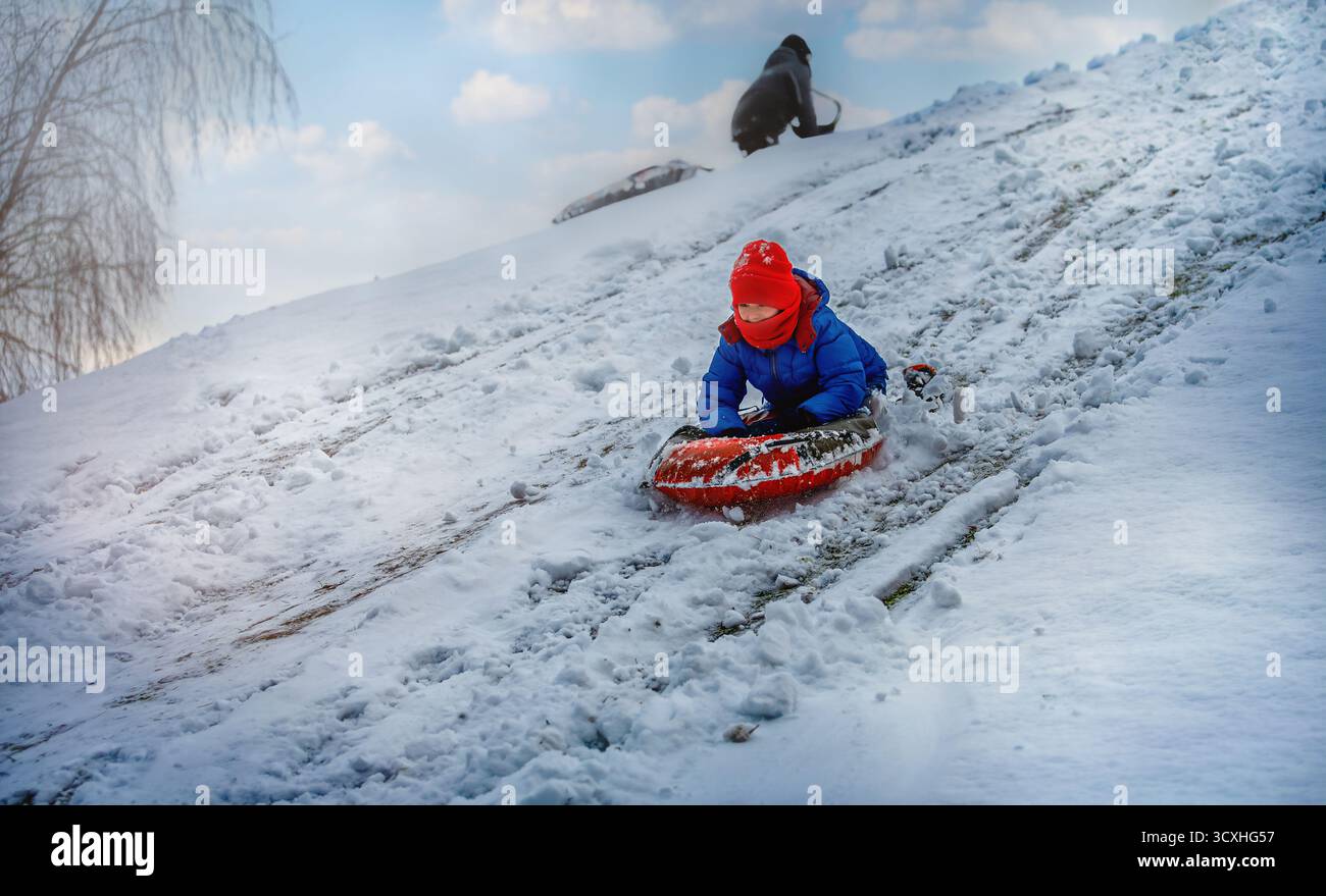 Ein Junge in hellen Winterkleidung rutscht auf einem aufblasbaren Schlauch in einem Park einen verschneiten Hügel hinunter. Die Szene fängt Bewegung, Freude und Outdoor-Spaß in der Sonne ein Stockfoto
