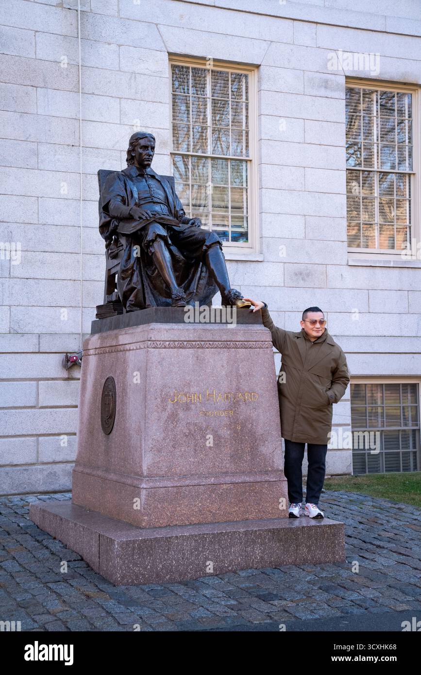 Ein Besucher posiert neben der John Harvard Statue in Harvard Yard und berührt den Bronzeschuh des Gründers, eine beliebte Tradition unter Campus-Touristen Stockfoto