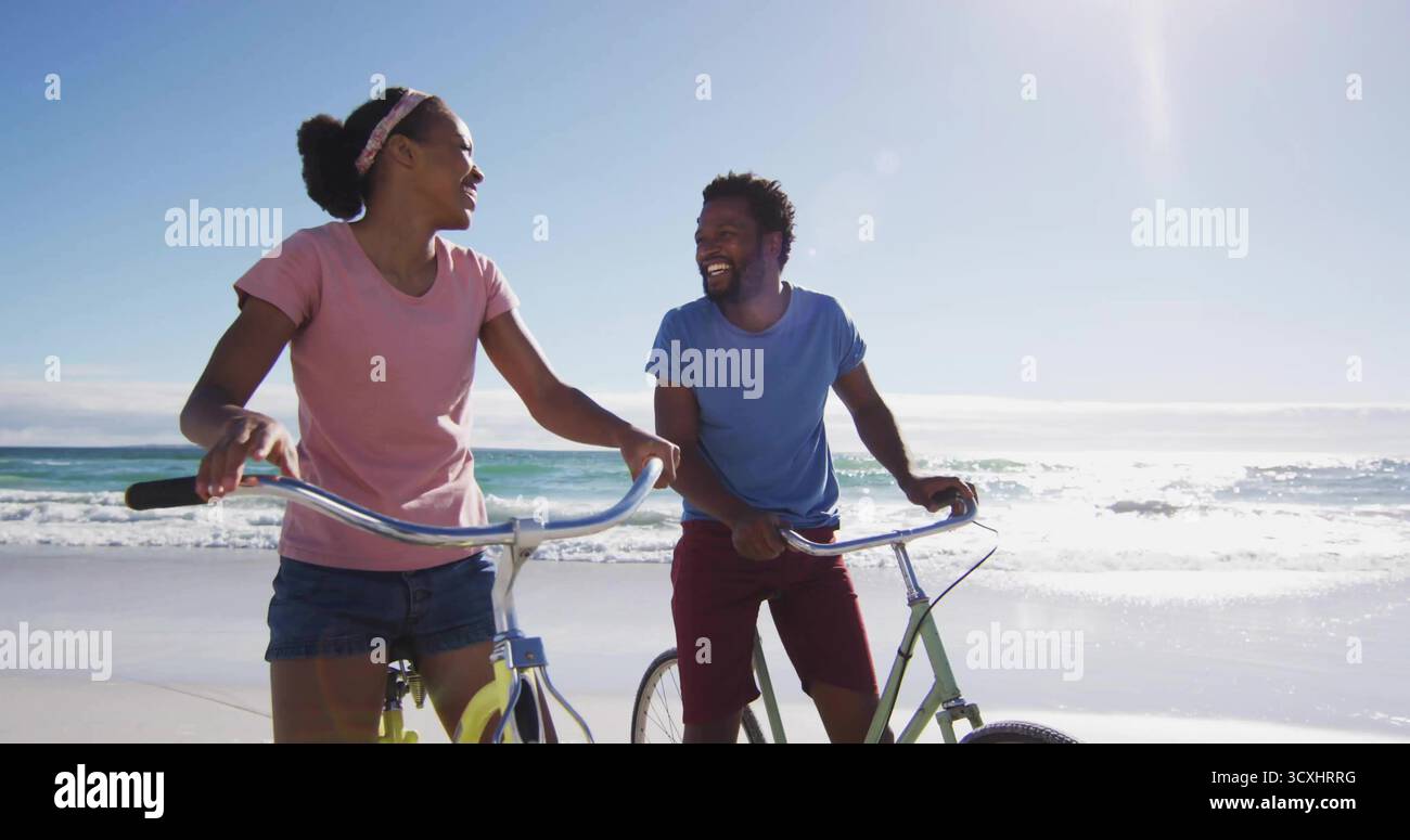 Lächelndes Paar, das unter Sonnenlicht an der Sandküste mit Fahrrädern unterwegs ist, T-Shirts und Shorts trägt Stockfoto