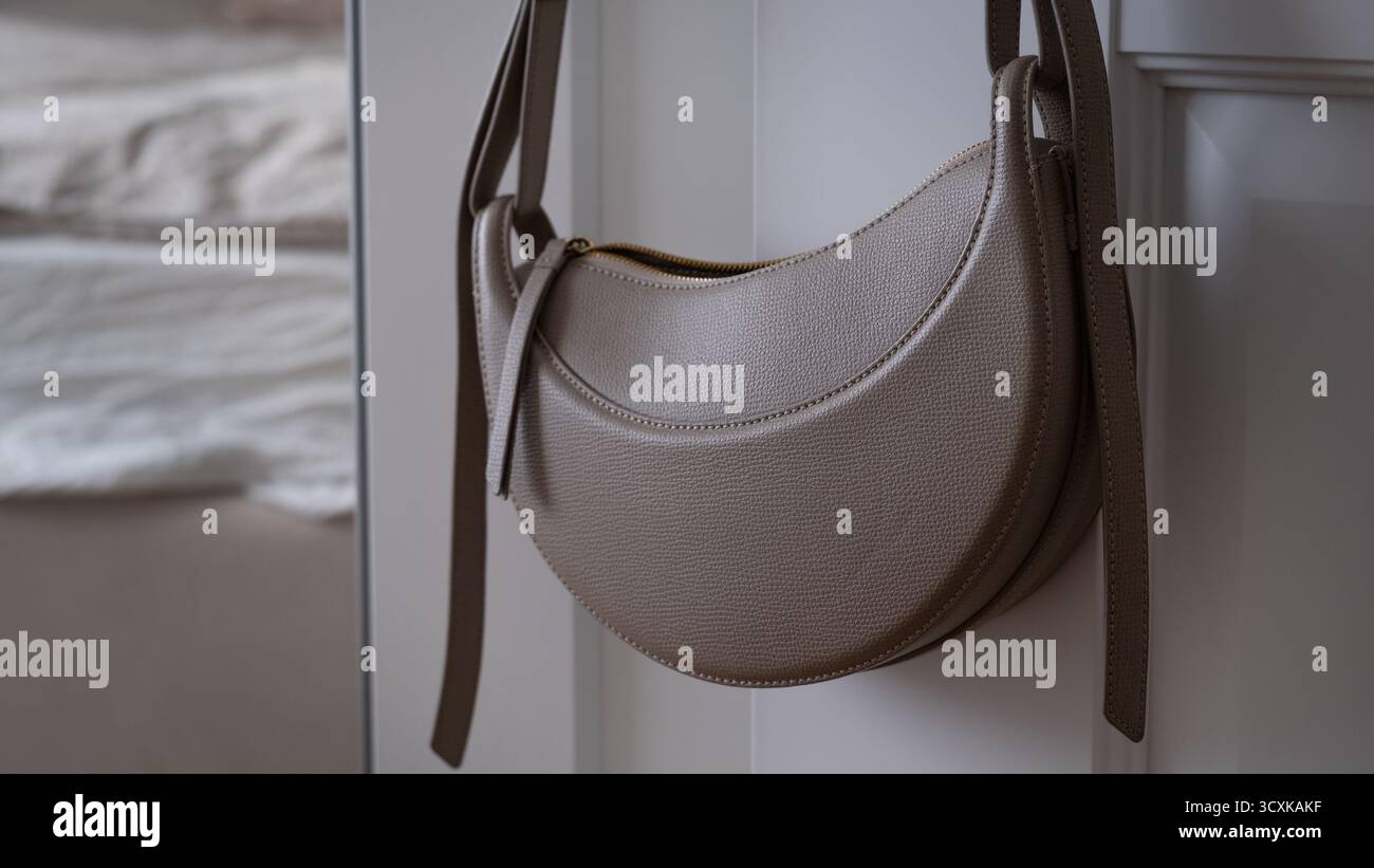 Stilvolle beige Handtasche aus halbmondförmigem Leder, die an der Schranktür in einem hellen Schlafzimmer hängt. Realistische, minimalistische Lifestyle-Szene mit natürlichem Licht und weicher Oberfläche Stockfoto