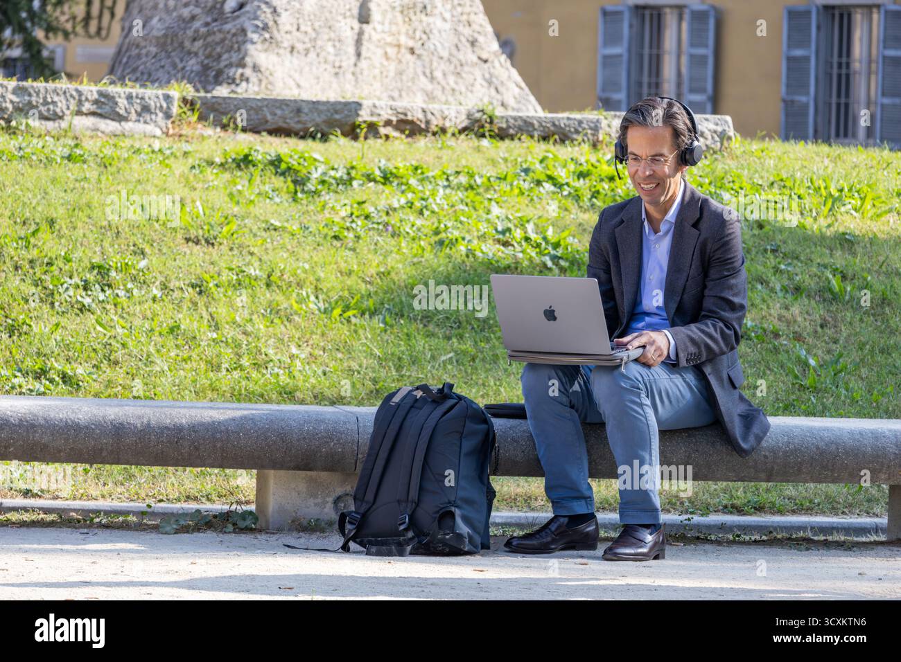 Ein lächelnder Geschäftsmann, der Kopfhörer trägt, arbeitet im Stadtpark bequem online und verbindet Technologie und Produktivität während eines Arbeitstages außerhalb des Büros. Stockfoto