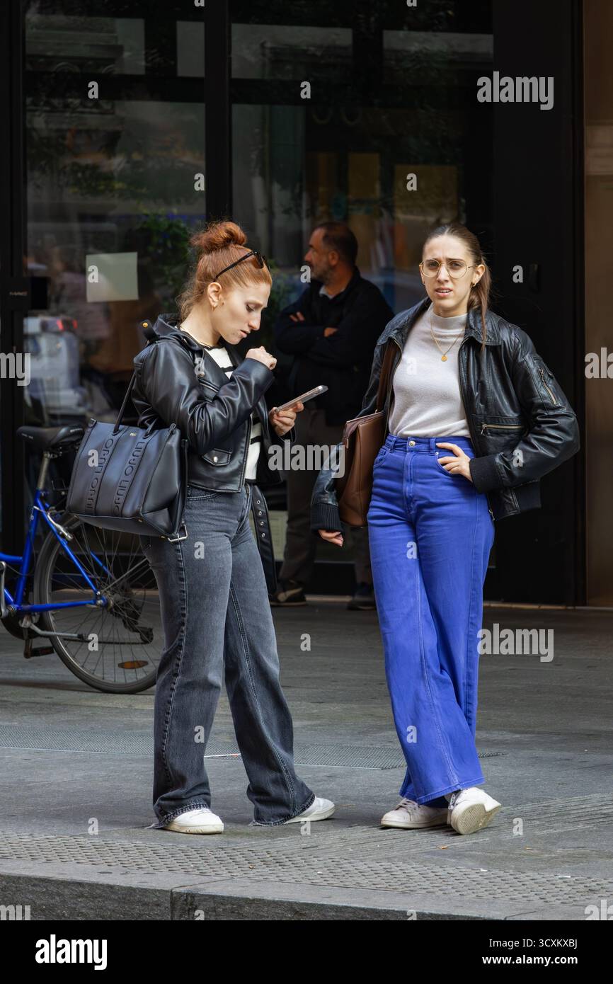 Zwei modische Frauen stehen auf der Straße der Stadt in trendigen Lederjacken, breiten Jeans, weißen Sneakers und modernem Freizeitstil. Stockfoto