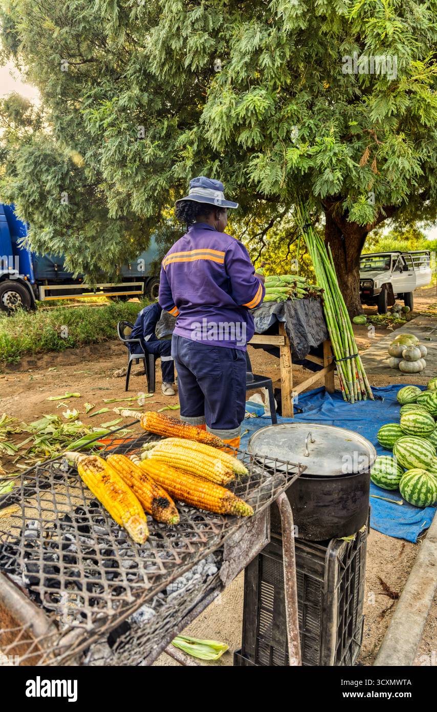 Eine afrikanische Straßenverkäuferin, die Mais, Kürbis und Wassermelone verkauft, am Straßenrand. Stockfoto
