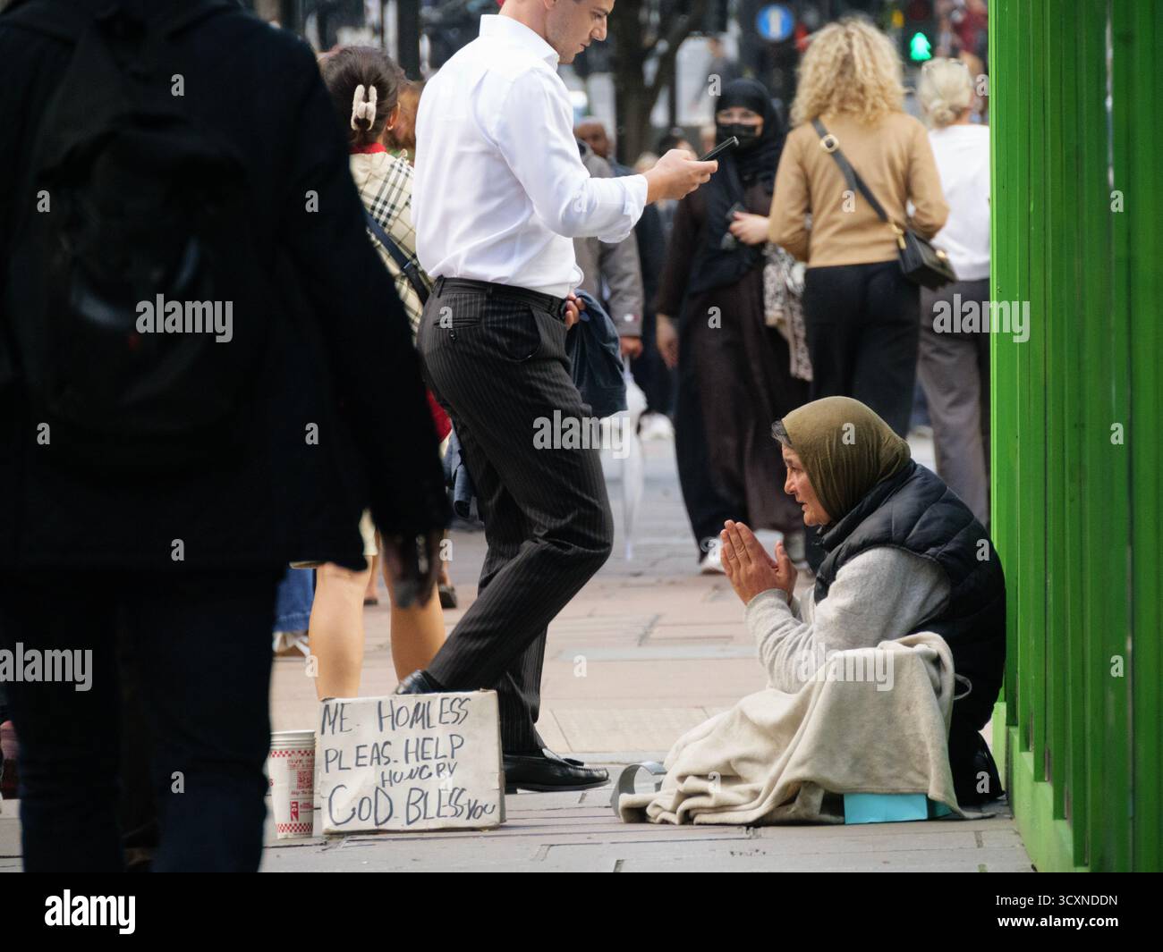 Eine ältere obdachlose Frau sitzt auf dem Bürgersteig und hält ein Schild mit der Aufschrift „Obdachlose Please help“, während die Käufer an der Oxford Street, London, Großbritannien, vorbeikommen Stockfoto Eine ältere obdachlose Frau sitzt auf dem Bürgersteig und hält ein Schild mit der Aufschrift „Obdachlose Please help“, während die Käufer an der Oxford Street, London, Großbritannien, vorbeikommen Stockfoto