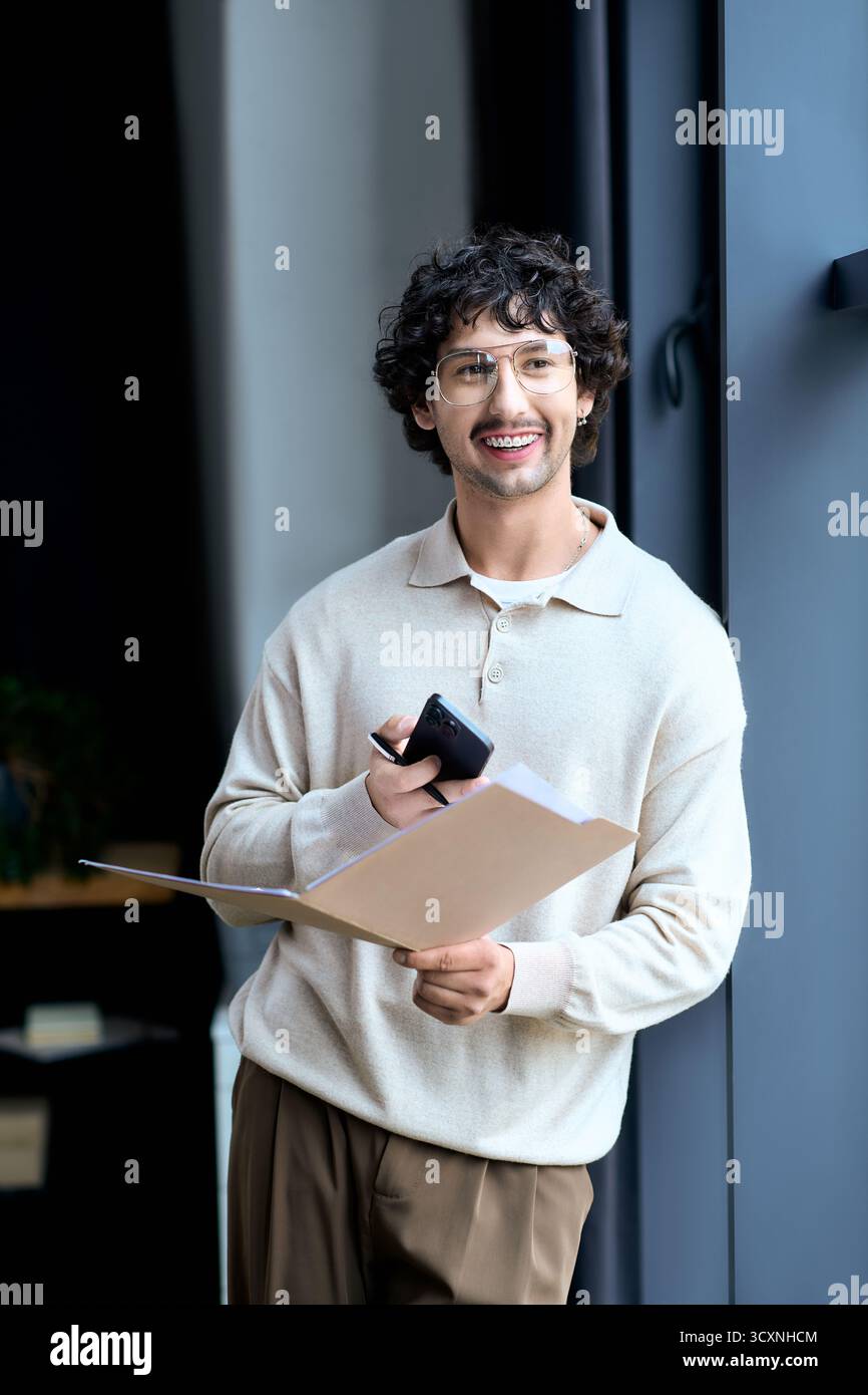 Der junge Mann genießt einen Moment in einem stilvollen Raum, hält eine Mappe und lächelt sein Handy an. Stockfoto