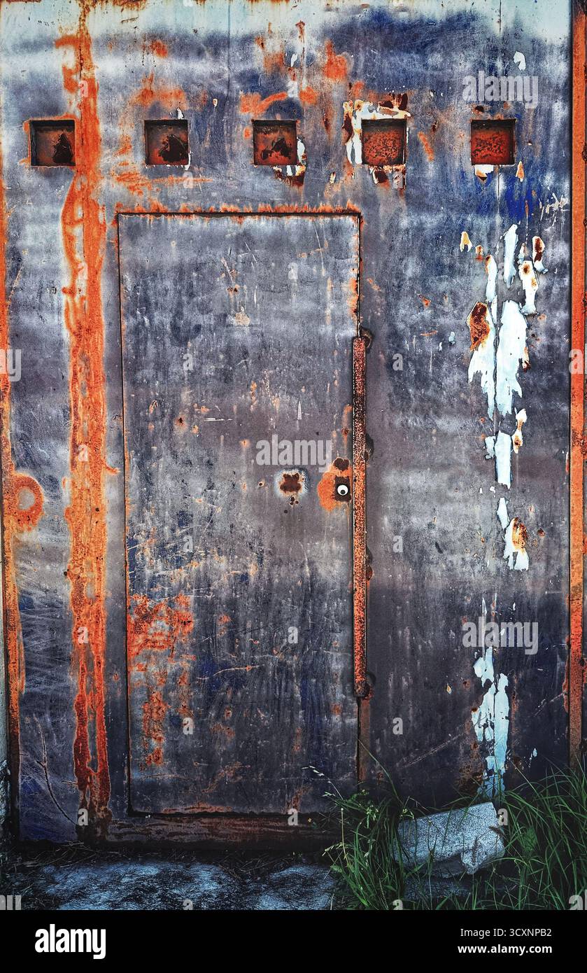 Verwitterte Metalltür mit Rost und abblätternder Farbe an der Wand eines Industriegebäudes, die Anzeichen von Verfall und Alter zeigt - Smartphone-aufgenommenes Stockfoto