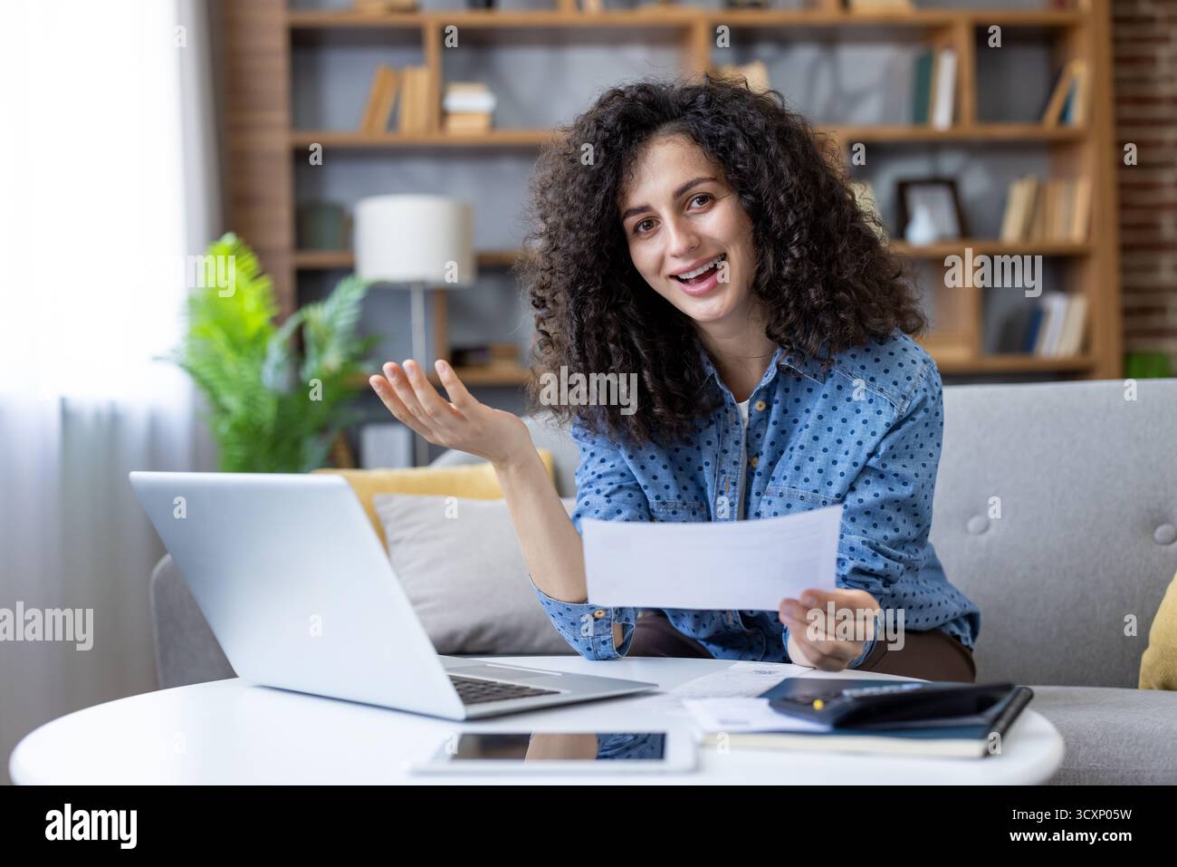 Glückliche junge Frau mit lockigem Haar, die sich um persönliche Finanzen kümmert und Rechnungen online bezahlt, in die Kamera schaut, während sie ein Dokument hält und auf einem Sofa sitzt Stockfoto
