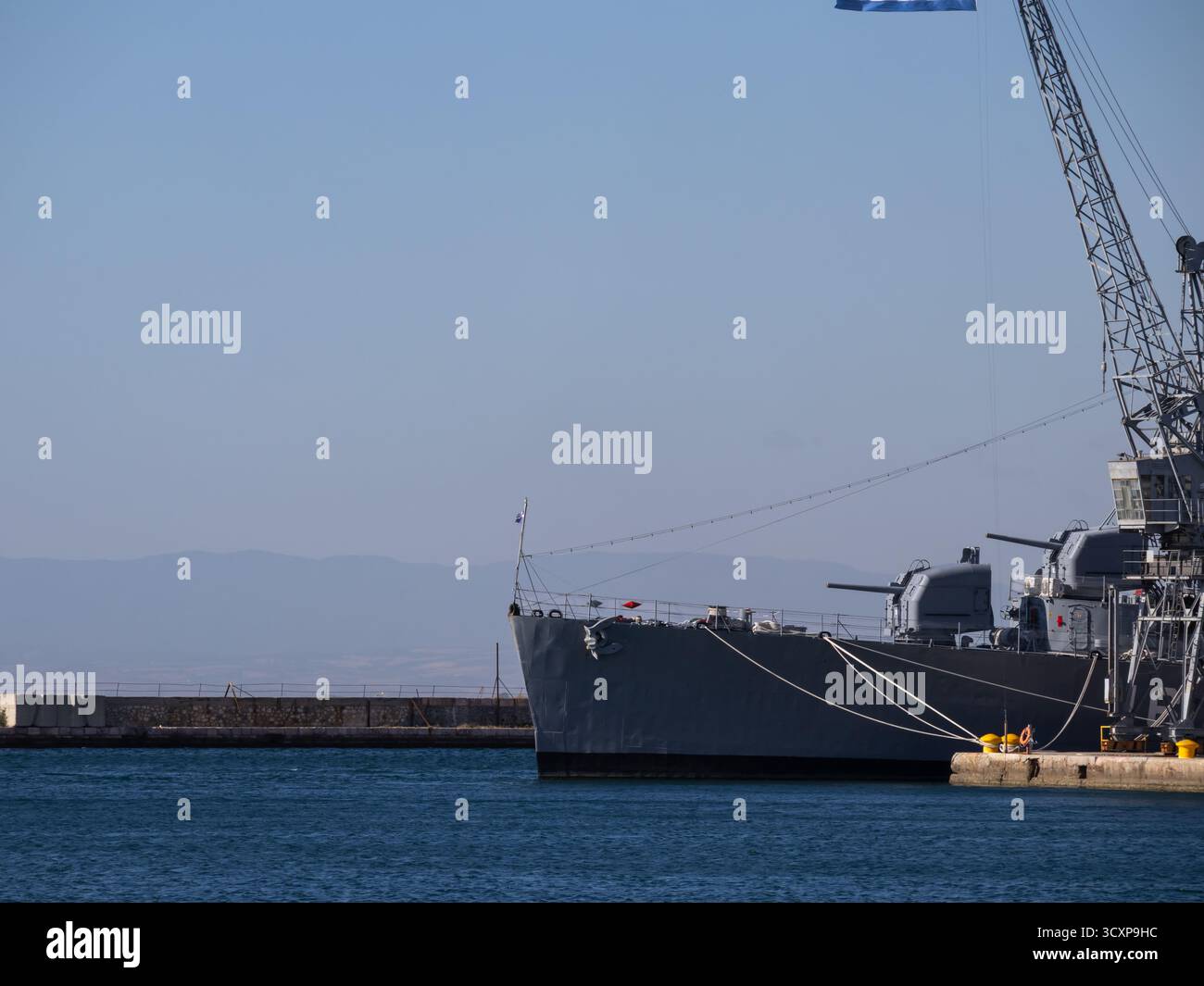 Der Bug des Marineschiffs legte an einem Hafen mit prominenten Artilleriegeschützen an, der neben einem großen Kran unter einem klaren blauen Himmel und einem entfernten, trüben Berg fest verankert war Stockfoto