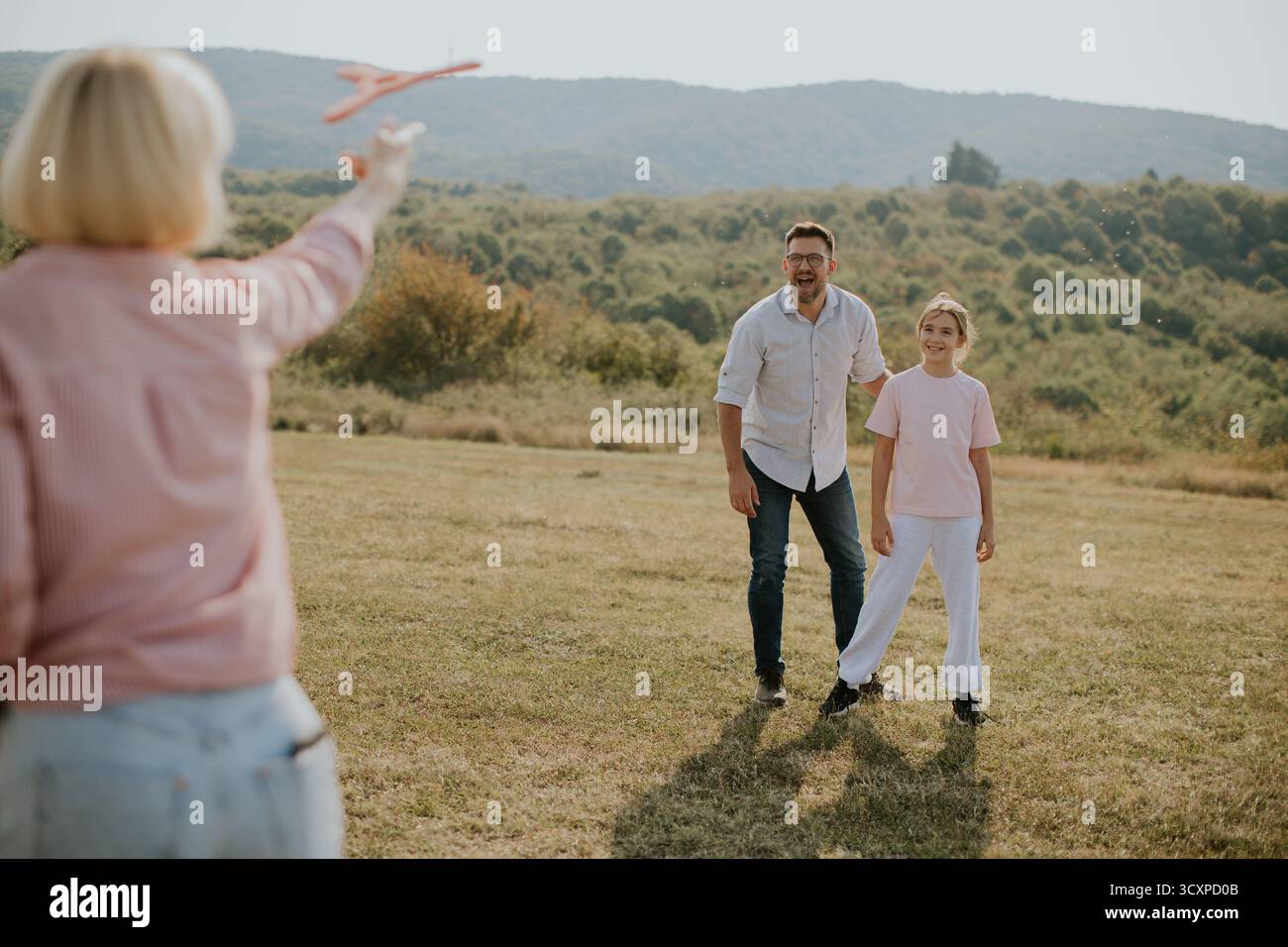 Eine Familie verbringt im Freien auf einem grasbewachsenen Feld eine schöne Zeit und wirft ein Spielzeugflugzeug in die Luft. Berge erheben sich im Hintergrund unter klarem Himmel, Crea Stockfoto