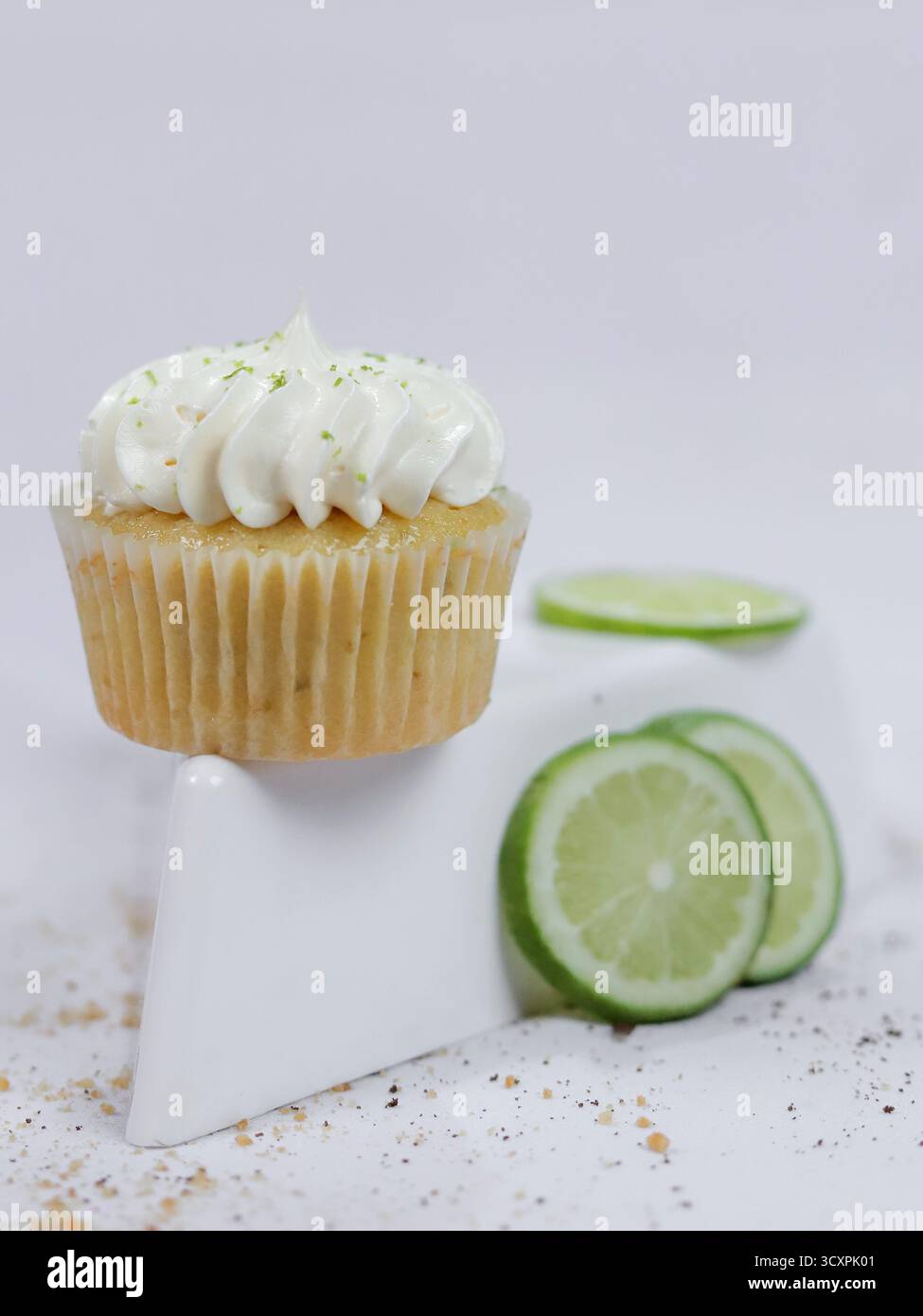 Ein frisch gebackener Zitronen-Cupcake mit cremigem Zuckerguss, der neben geschnittenen Limetten auf einer sauberen weißen Oberfläche platziert wird Stockfoto