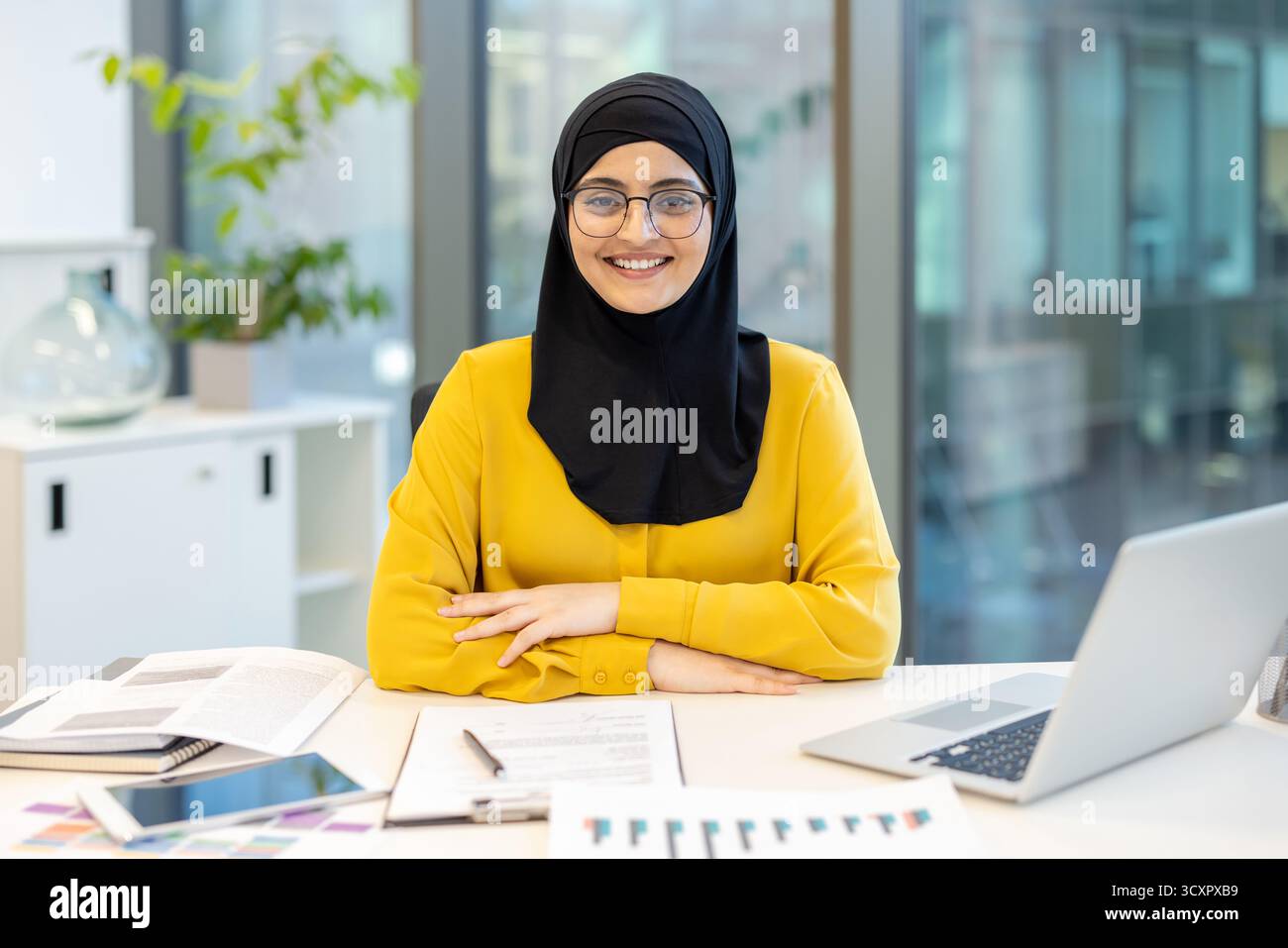 Junge muslimische Geschäftsfrau in Hijab und Brille, die positiv lächelt, während sie an einem Schreibtisch in einem zeitgenössischen Büroumfeld sitzt und Professionsio repräsentiert Stockfoto Junge muslimische Geschäftsfrau in Hijab und Brille, die positiv lächelt, während sie an einem Schreibtisch in einem zeitgenössischen Büroumfeld sitzt und Professionsio repräsentiert Stockfoto