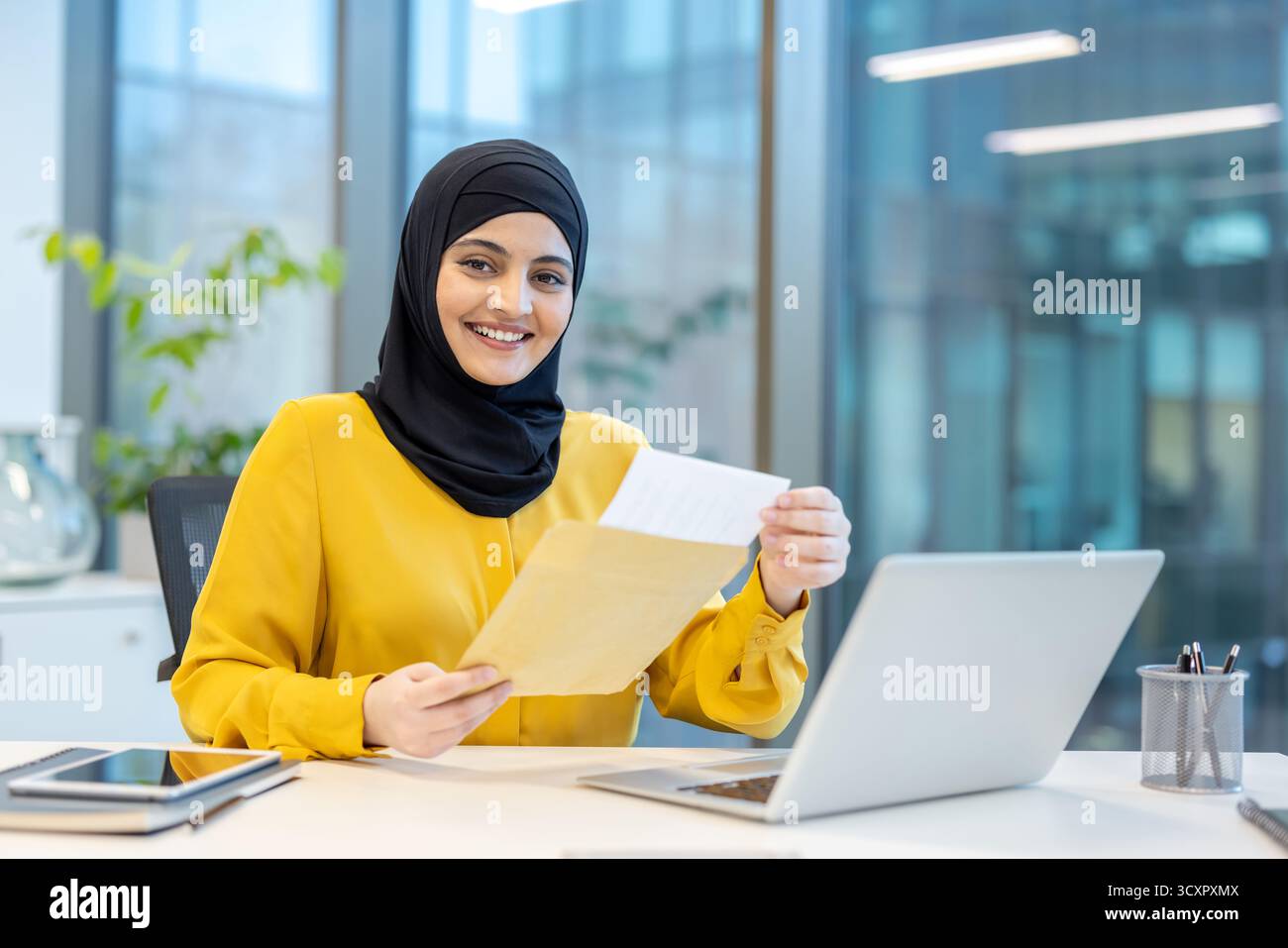 Muslimische Geschäftsfrau in einem Hijab, lächelt in die Kamera, während sie einen Umschlag mit einem Dokument öffnet und mit A la an einem modernen Schreibtisch sitzt Stockfoto Muslimische Geschäftsfrau in einem Hijab, lächelt in die Kamera, während sie einen Umschlag mit einem Dokument öffnet und mit A la an einem modernen Schreibtisch sitzt Stockfoto