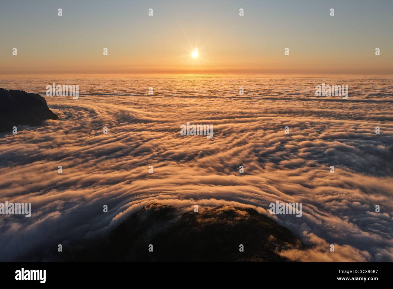 Die Sonne geht über einer dicken Wolkenschicht auf, die einem Ozean ähnelt, und bietet einen atemberaubenden Blick auf die Berge, die während des Sonnenaufgangs durch den goldenen Nebel ragen Stockfoto