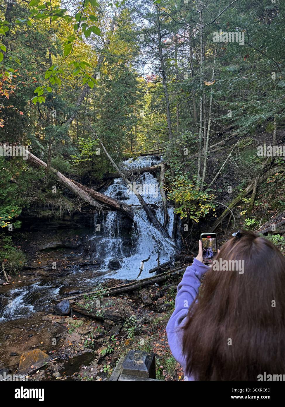 Der Rücken einer Frau, die ein Foto von Wagner Falls macht, die über Felsen und umgestürzte Bäume einen Hügel hinunterfallen, im Wald in Munising, Michigan, USA - Smartphone-aufgenommenes Stockfoto