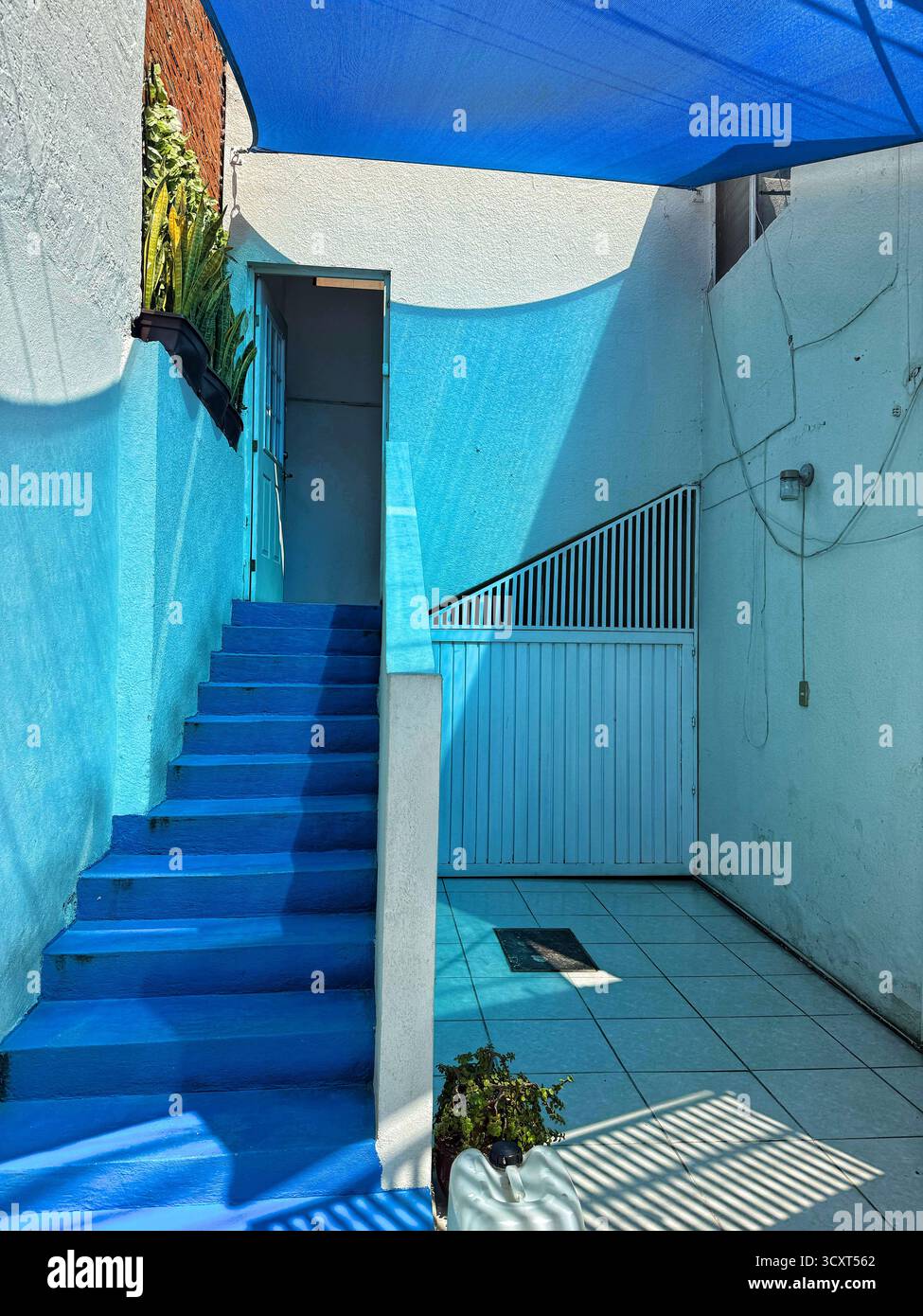 Eine hellblaue Treppe führt zu einem von weißen Wänden umgebenen Innenhof, dessen Sonnenlicht scharfe Schatten über den Boden wirft - Smartphone-aufgenommenes Stockfoto