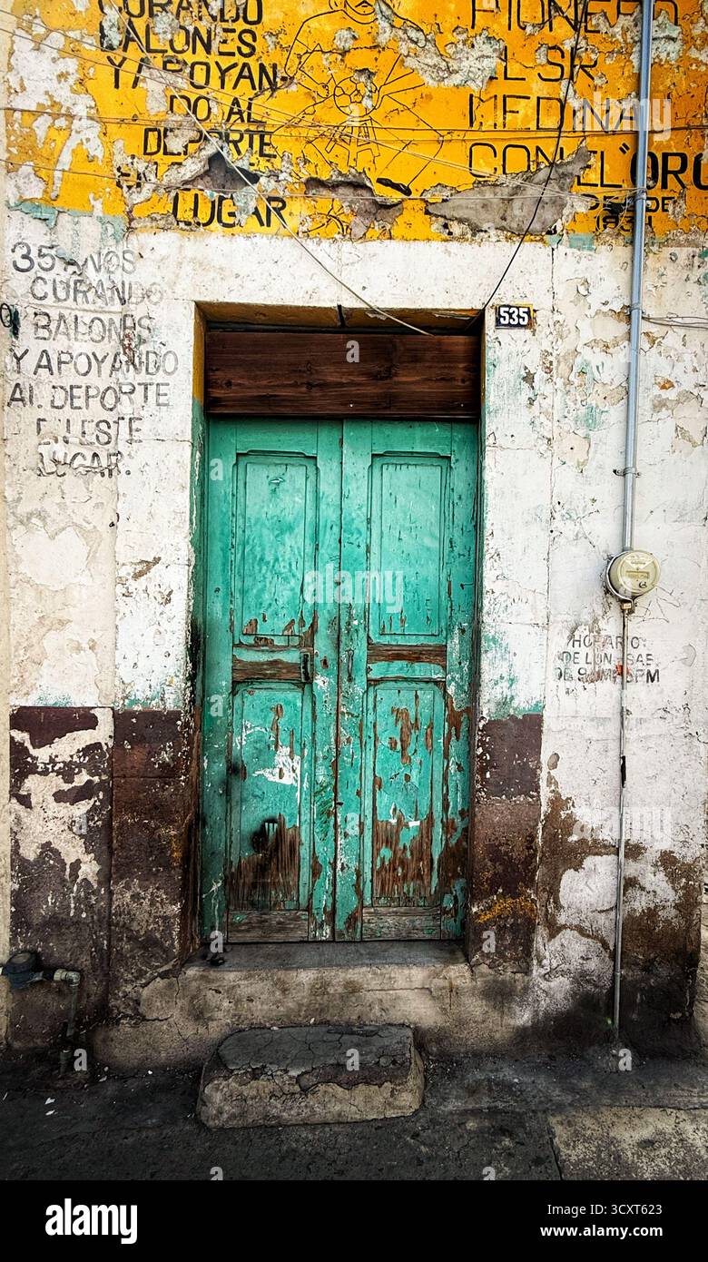 Eine verwitterte türkisfarbene Holztür mit abblätternder Farbe in einer alten Wand, die mit verblassten Werbespots und Graffiti bedeckt ist. Stockfoto