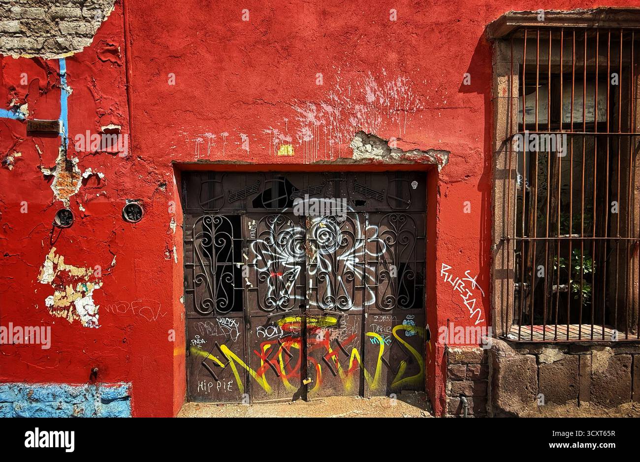 Eine verwitterte rote Wand mit lebendigen Graffiti über einem Metalltor, die urbane Kunst in einem rustikalen Viertel zeigt Stockfoto