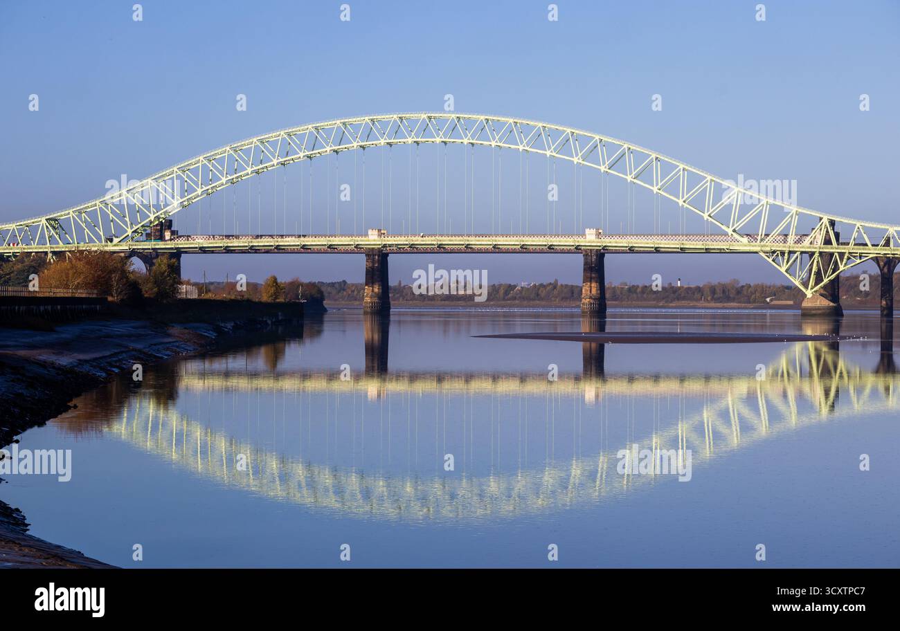 Die Silver Jubilee Bridge überspannt den Fluss Mersey zwischen Runcorn und Widnes, die sich bei Sonnenaufgang in ruhigem Wasser spiegelt Stockfoto