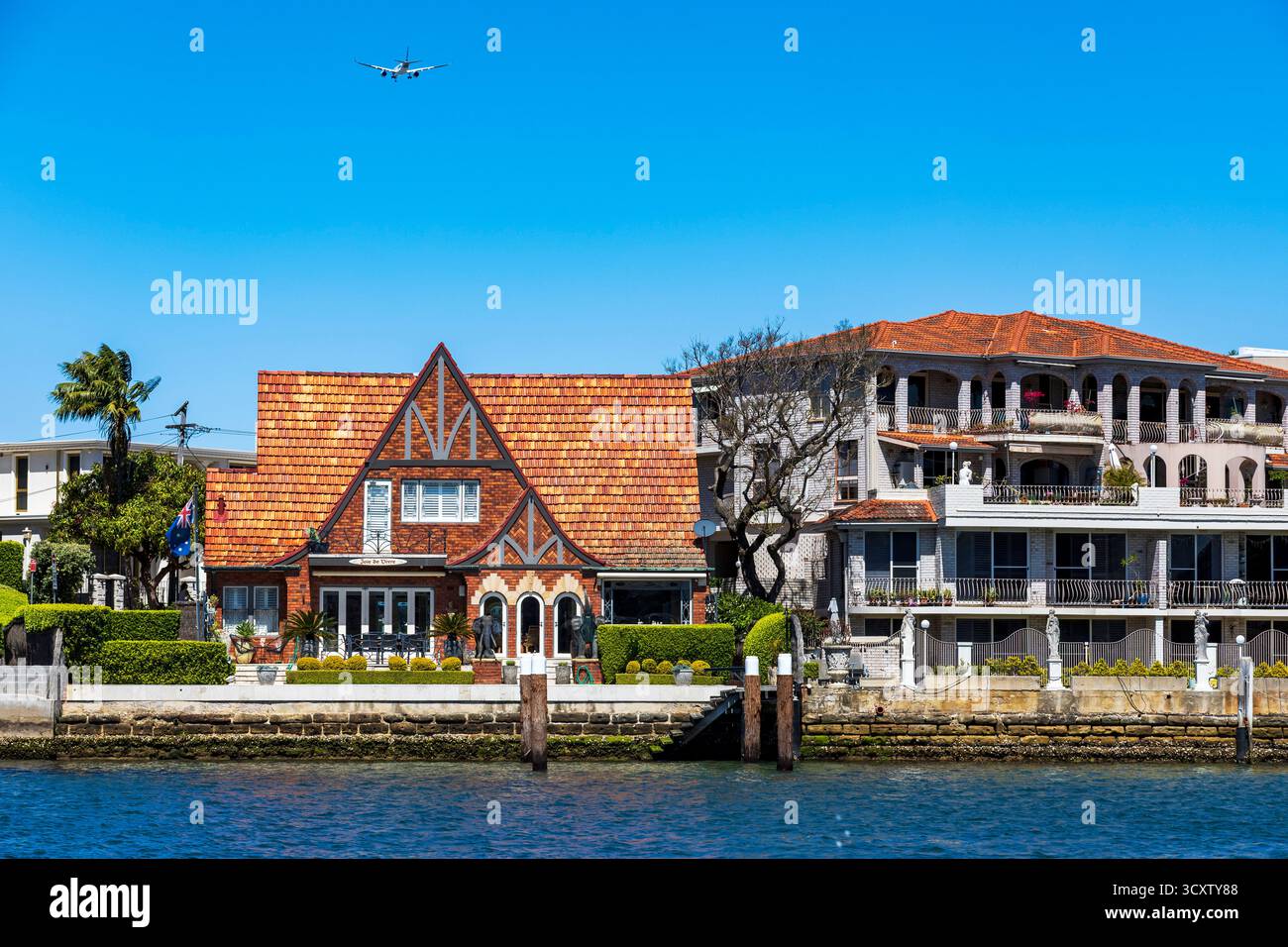 Häuser am Wasser in einem lebendigen Viertel mit klarem blauen Himmel und einem Flugzeug im Flug, Drummoyne Wharf, Parramatta River, Sydney Harbour, Stockfoto