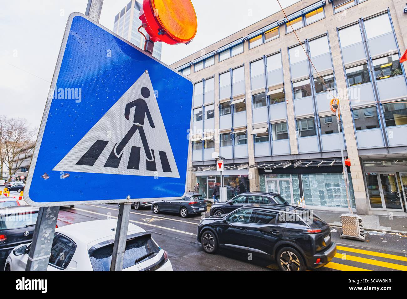Ein Fußgängerschild mit einer orangefarbenen Warnleuchte darüber, das sich vor einer belebten Stadtstraße und einem bewölkten Himmel befindet Stockfoto