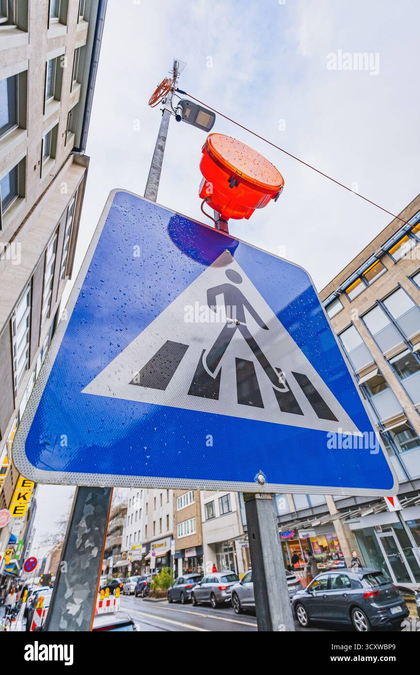 Ein Fußgängerschild mit einer orangefarbenen Warnleuchte darüber, das sich vor einer belebten Stadtstraße und einem bewölkten Himmel befindet Stockfoto