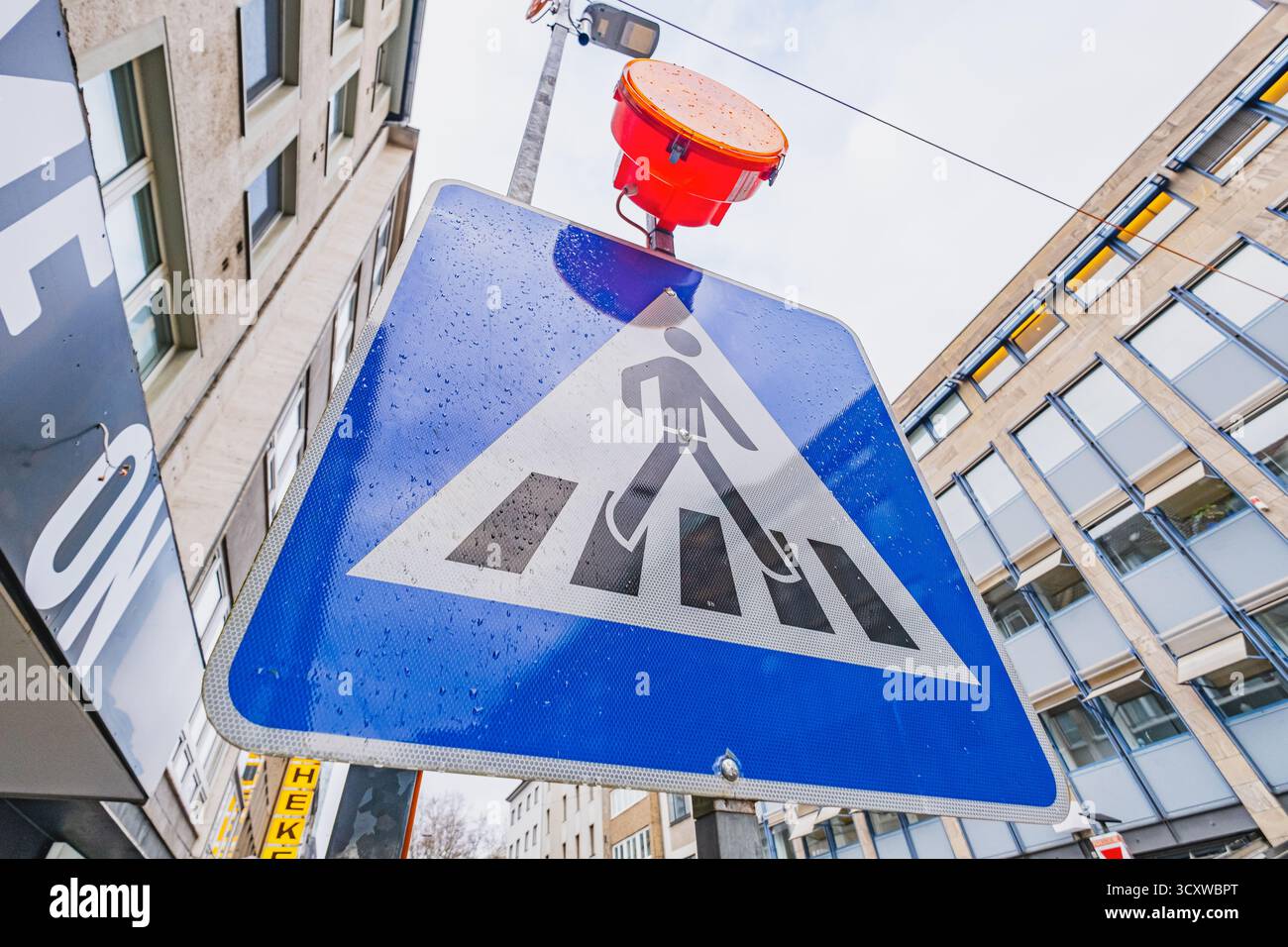 Ein Fußgängerschild mit einer orangefarbenen Warnleuchte darüber, das sich vor einer belebten Stadtstraße und einem bewölkten Himmel befindet Stockfoto