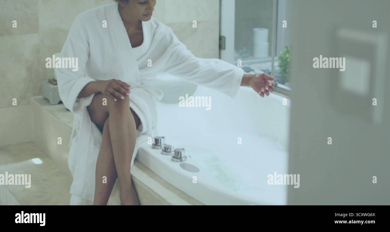 Frau in weißem Bademantel taucht Hand in schäumendes Wasser in Marmorbadewanne, mit grauem Kasten, Fenster Stockfoto