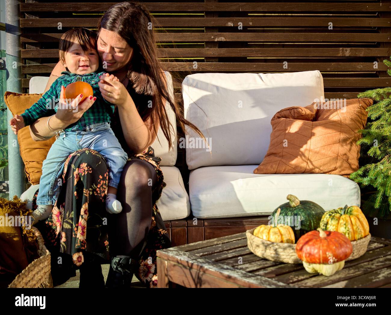 Gemütlicher Herbstmoment: Person oder Mutter hält ein Baby mit Kürbis auf einem Sofa im Freien, umgeben von Kürbissen und herbstlicher Atmosphäre. Konzept der Verklebung Stockfoto