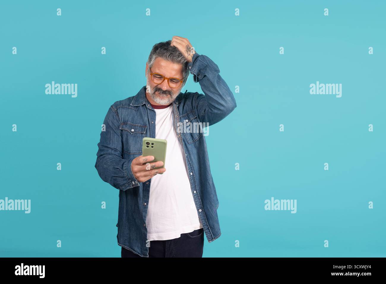 Reifer Mann kratzt den Kopf, während er auf seinen Handy-Bildschirm blickt und Verwirrung ausdrückt Stockfoto